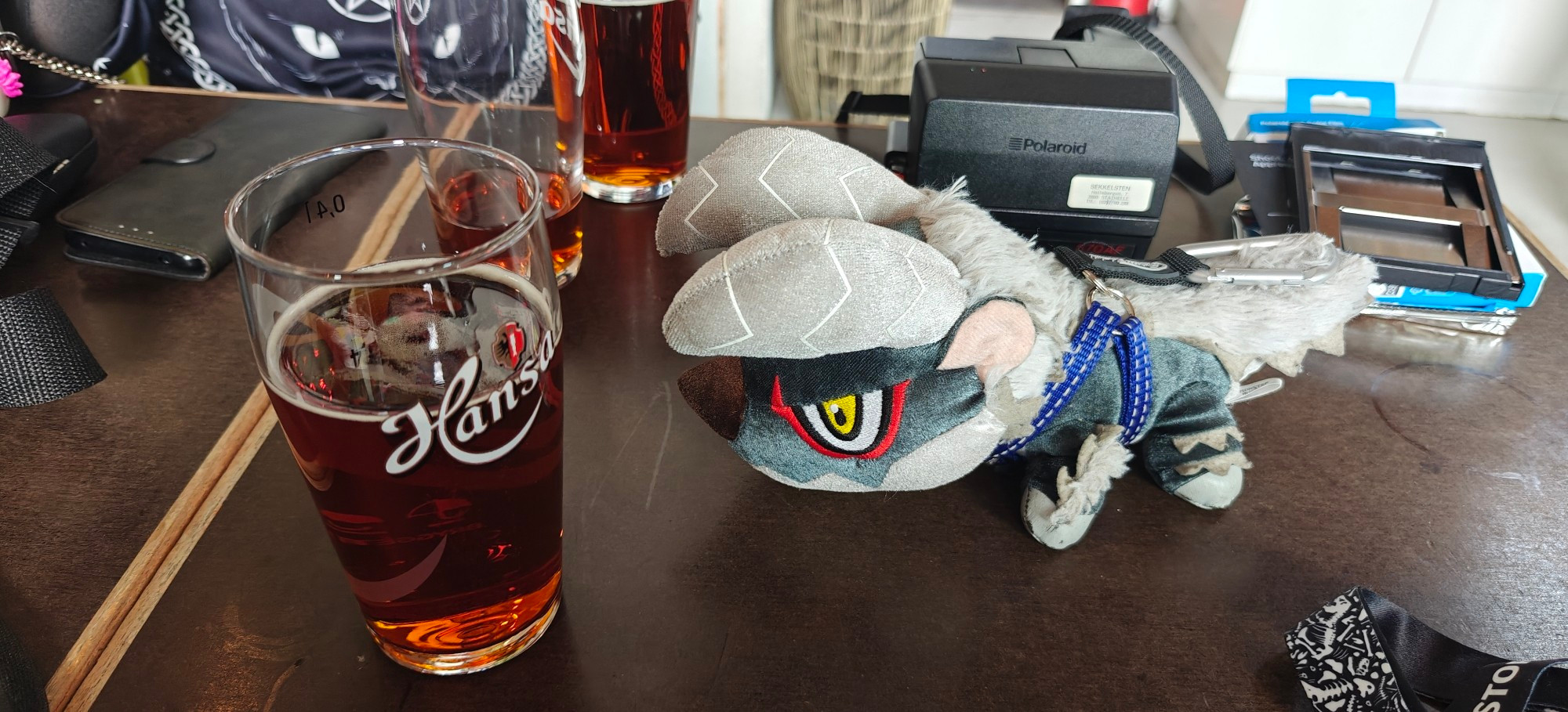 Deformed Stygian Zinogre plushie looking at a glass of beer. There's a Polaroid 670AF behind it, and the glass is half full of Bayer