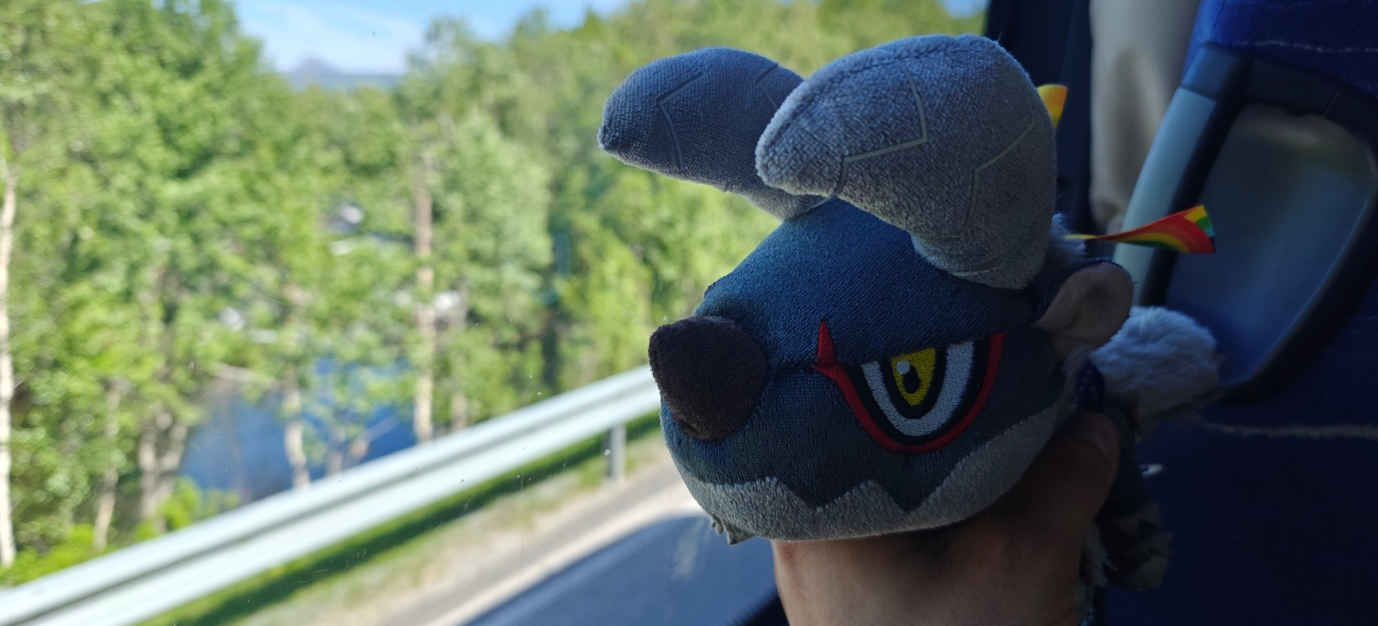 Picture of a deformed Stygian Zinogre plushie looking out the window of a bus that's driving down a lightly forested countryside road