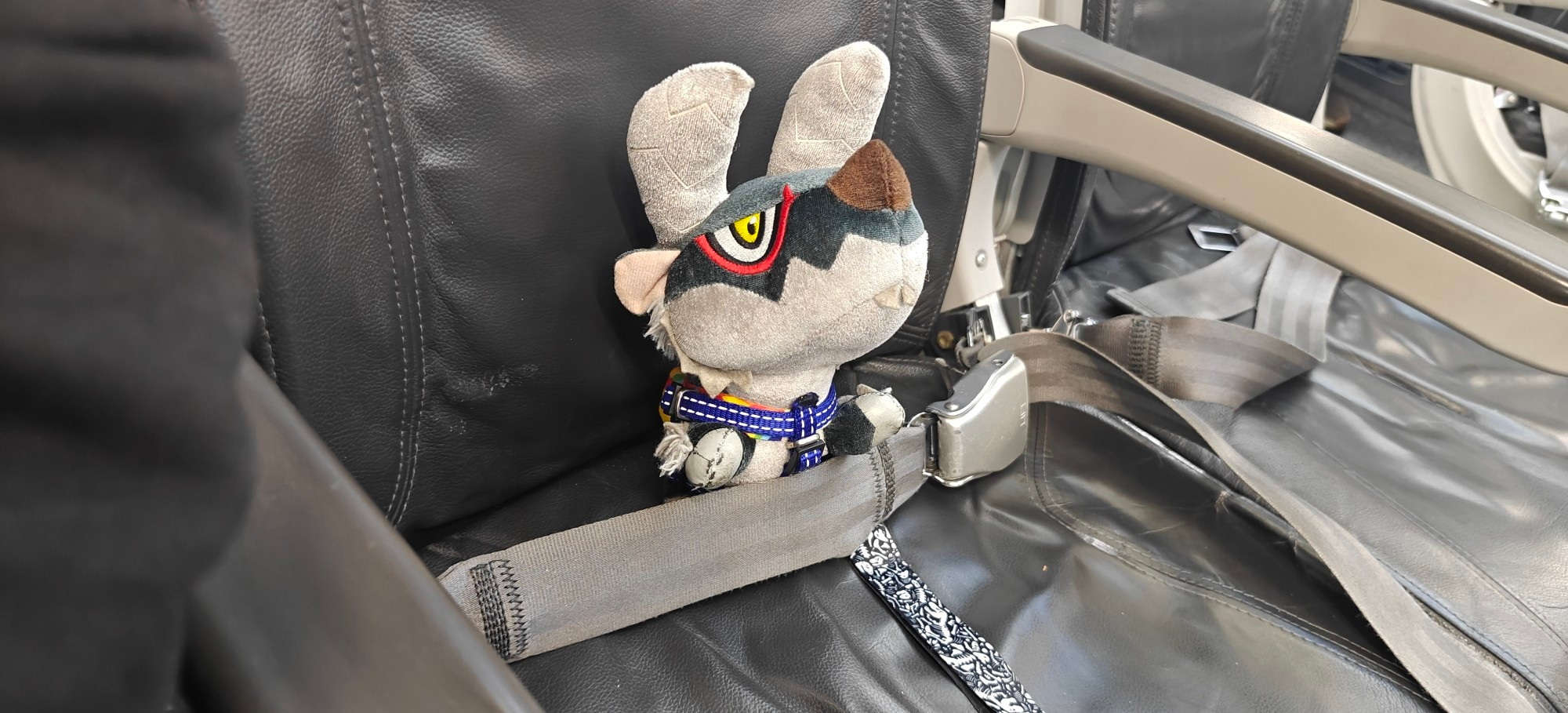 Deformed Stygian Zinogre plushie in a plane seat, strapped in like you should be when flying