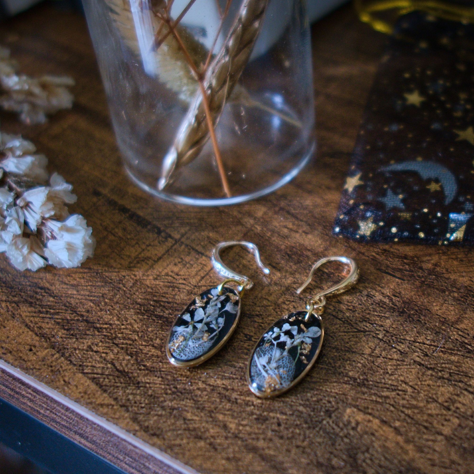 Boucles d'oreilles ORACLE d'Ortilys aux contours dorés, composés de pétales d'hortensias, fleur de Carotte, gypsophiles et de feuilles d'or sur fond noir