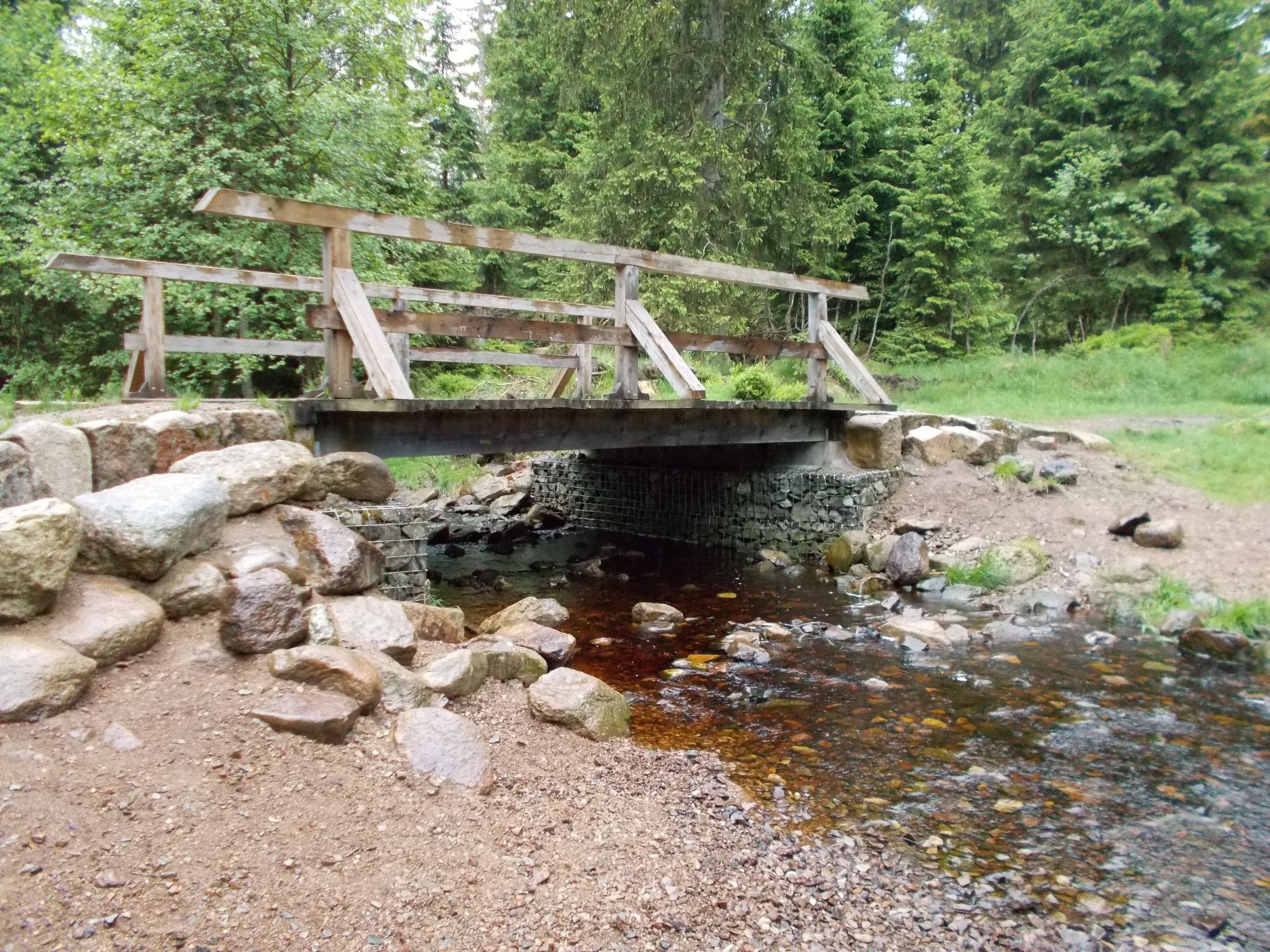 Eine Holzbrücke mitten im Wald führt über einen kleinen Fluss, der in einem Bett aus Geröll und Kies fließt. Das Wasser ist leicht braunrot vom umgebenden Moor. Die Ufer an der Brücke sind mit größeren Feldsteinen und Kies befestigt.