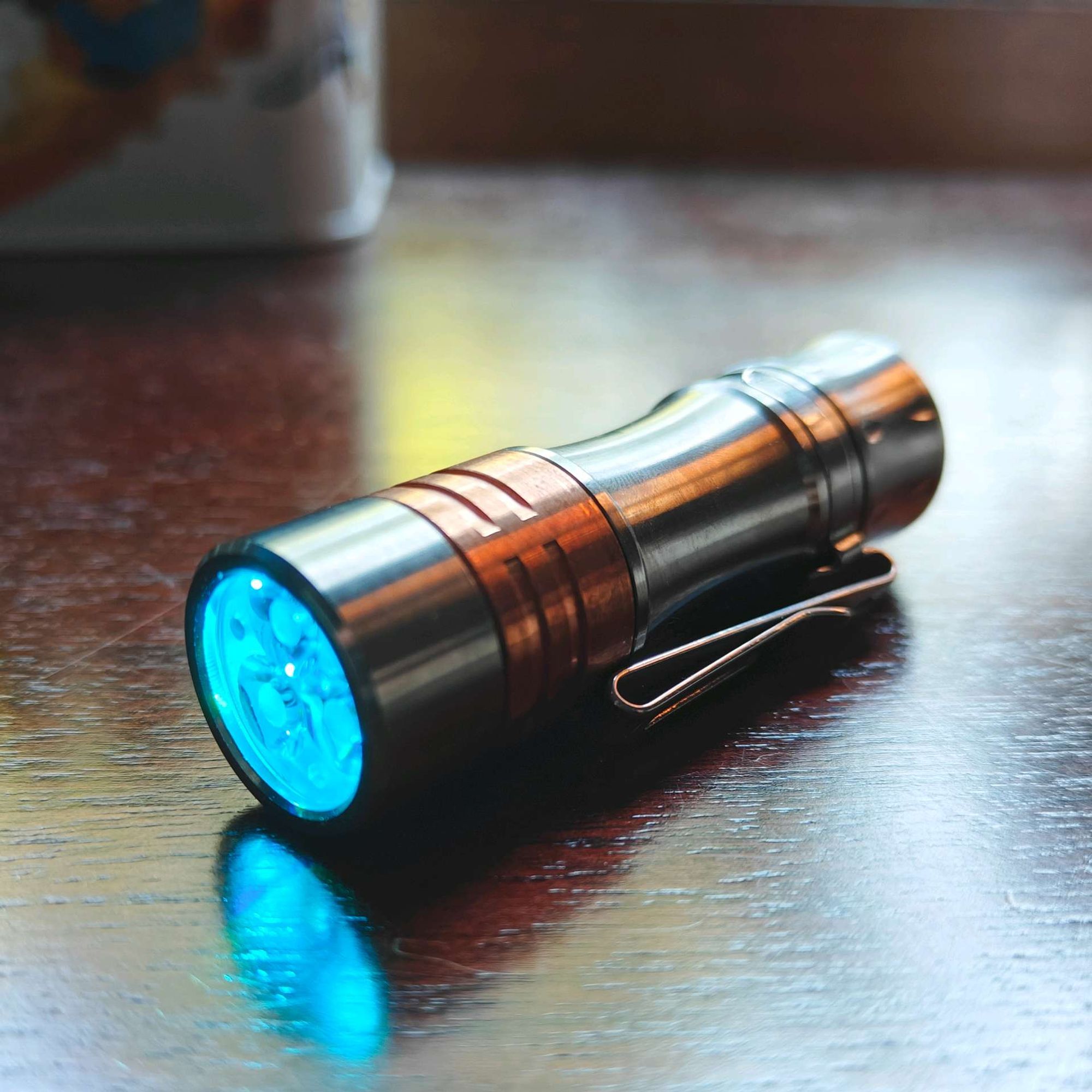 A titanium and copper mini flashlight sitting on a wooden desktop. The led lights have a cool blue glow. 