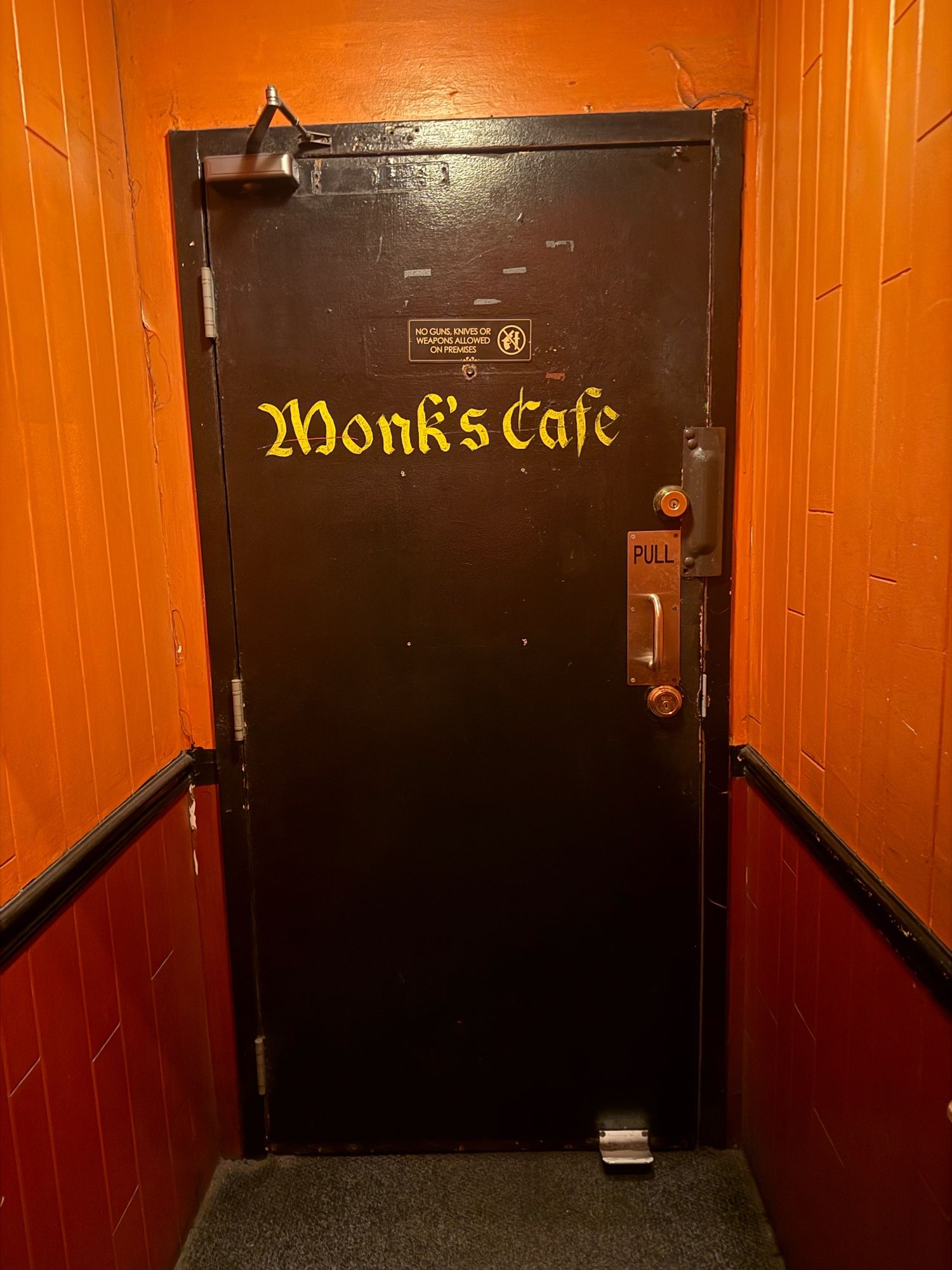 A black door at the end of a orange and red tile hallway with the words “Monk’s Cafe” painted on the door