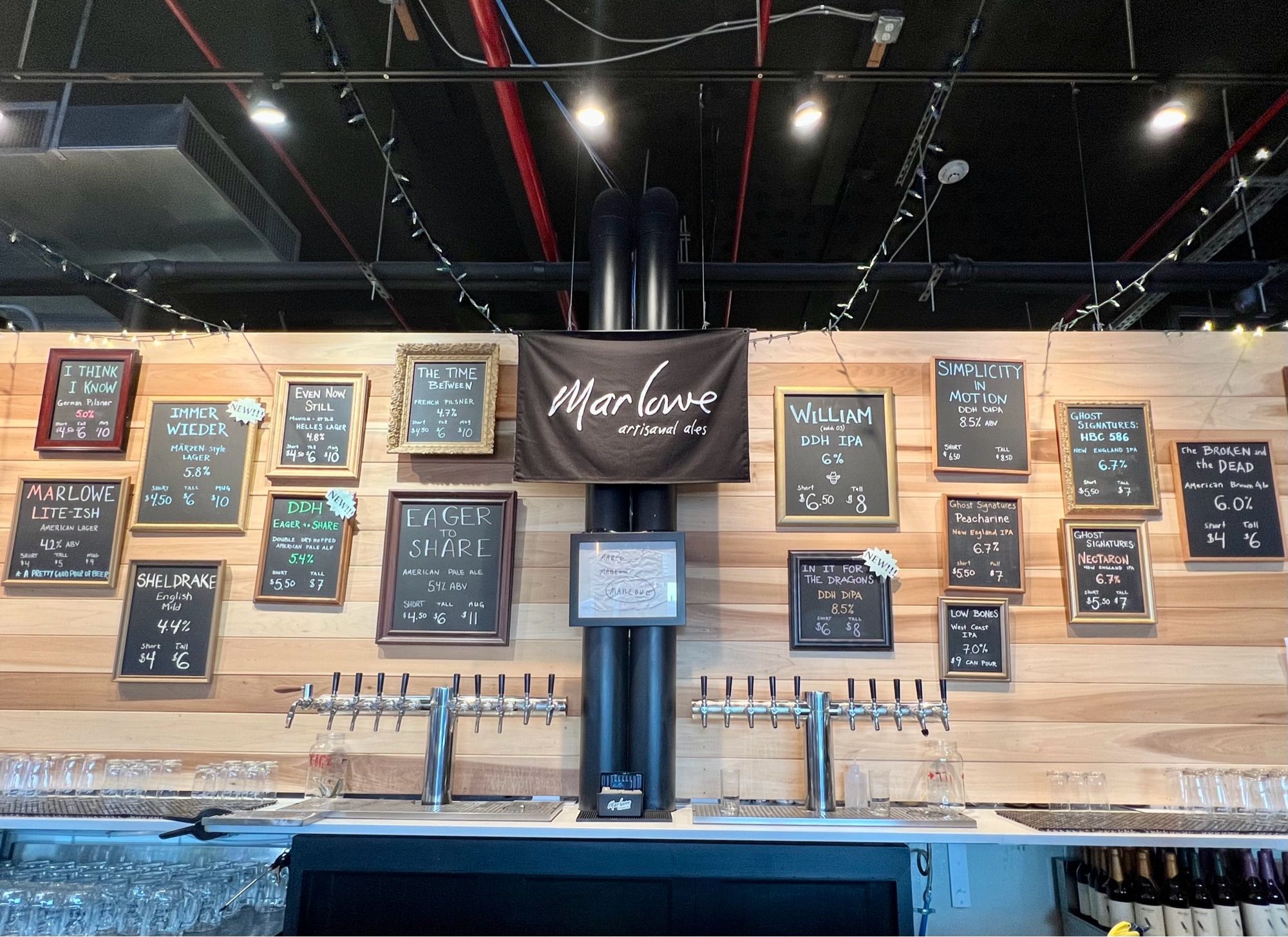 A brewery taproom’s back bar, with beer taps in the foreground and a wall with framed chalkboards displaying beer menu options in the background.