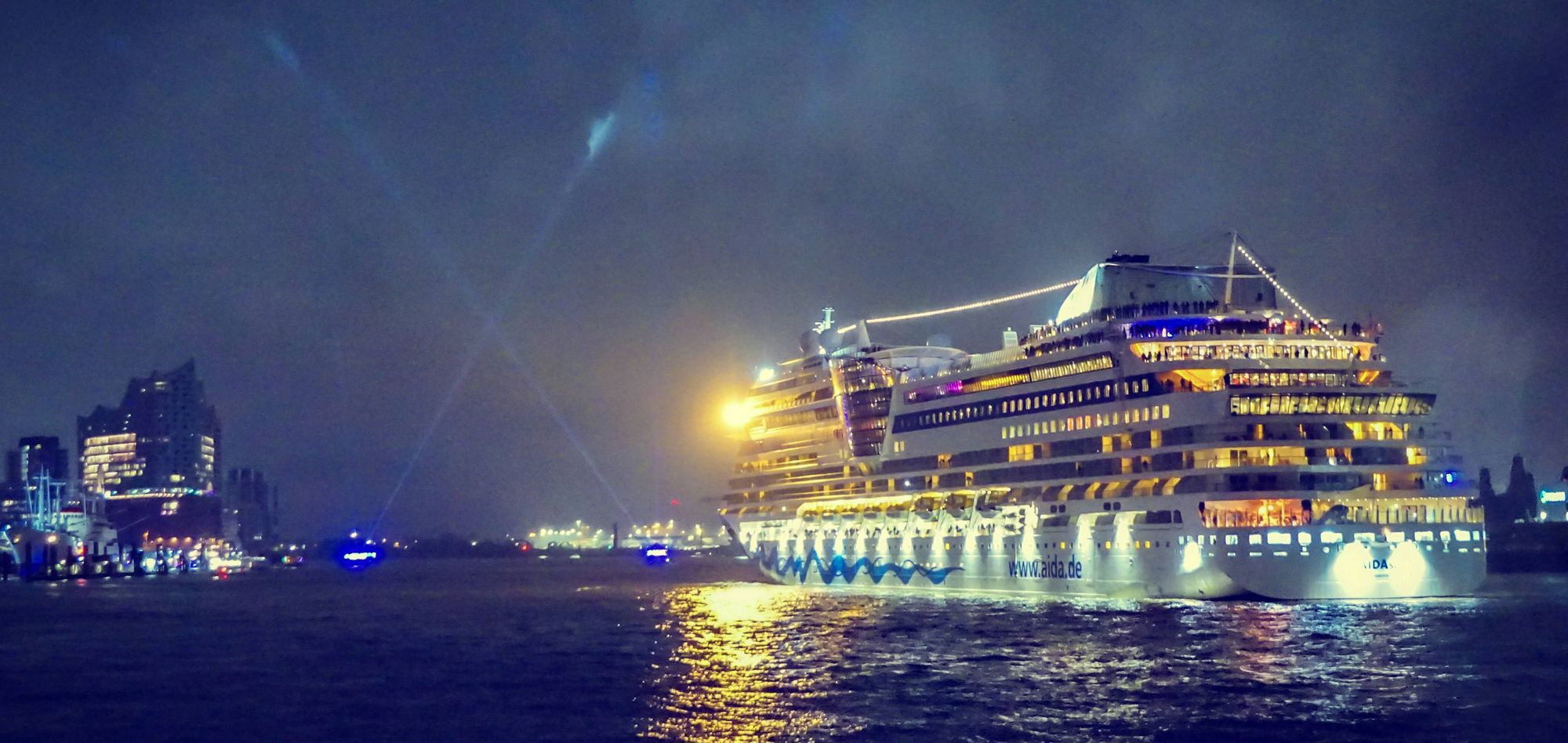 Die beleuchte AIDAsol fährt die Elbe hinauf. Im Hintergrund links die Elbphilharmonie, rechts davon zwei Schlepper, die mit Scheinwerfern ein X in den diesigen Nachthimmel leuchten.