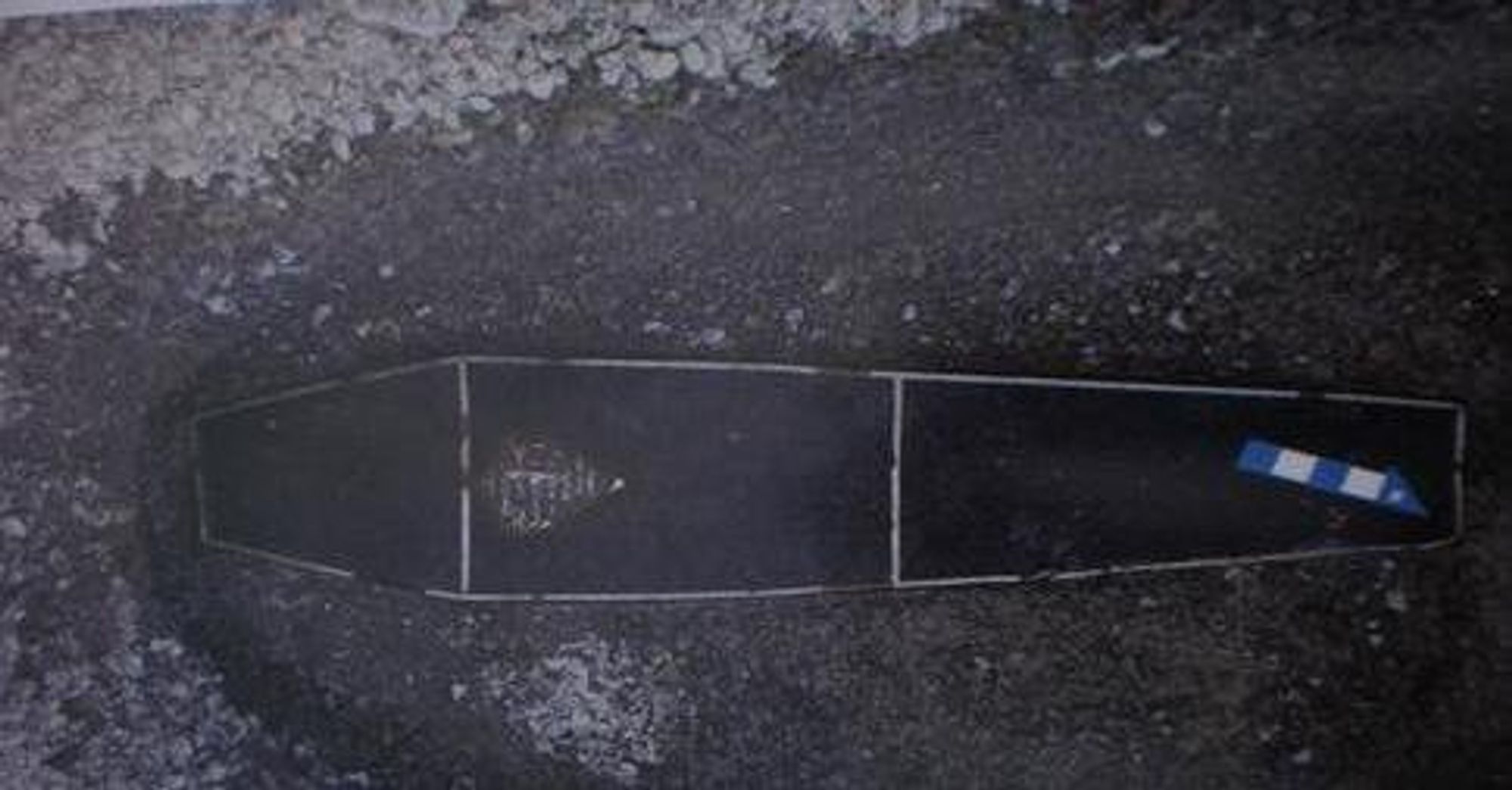 A short, narrow coffin sitting in a hole. The coffin is covered in dark blue fabric with white lines along the sides and crossing through the middle. A plaque is attached near the head of the coffin and a blue and white arrow lays at the foot.