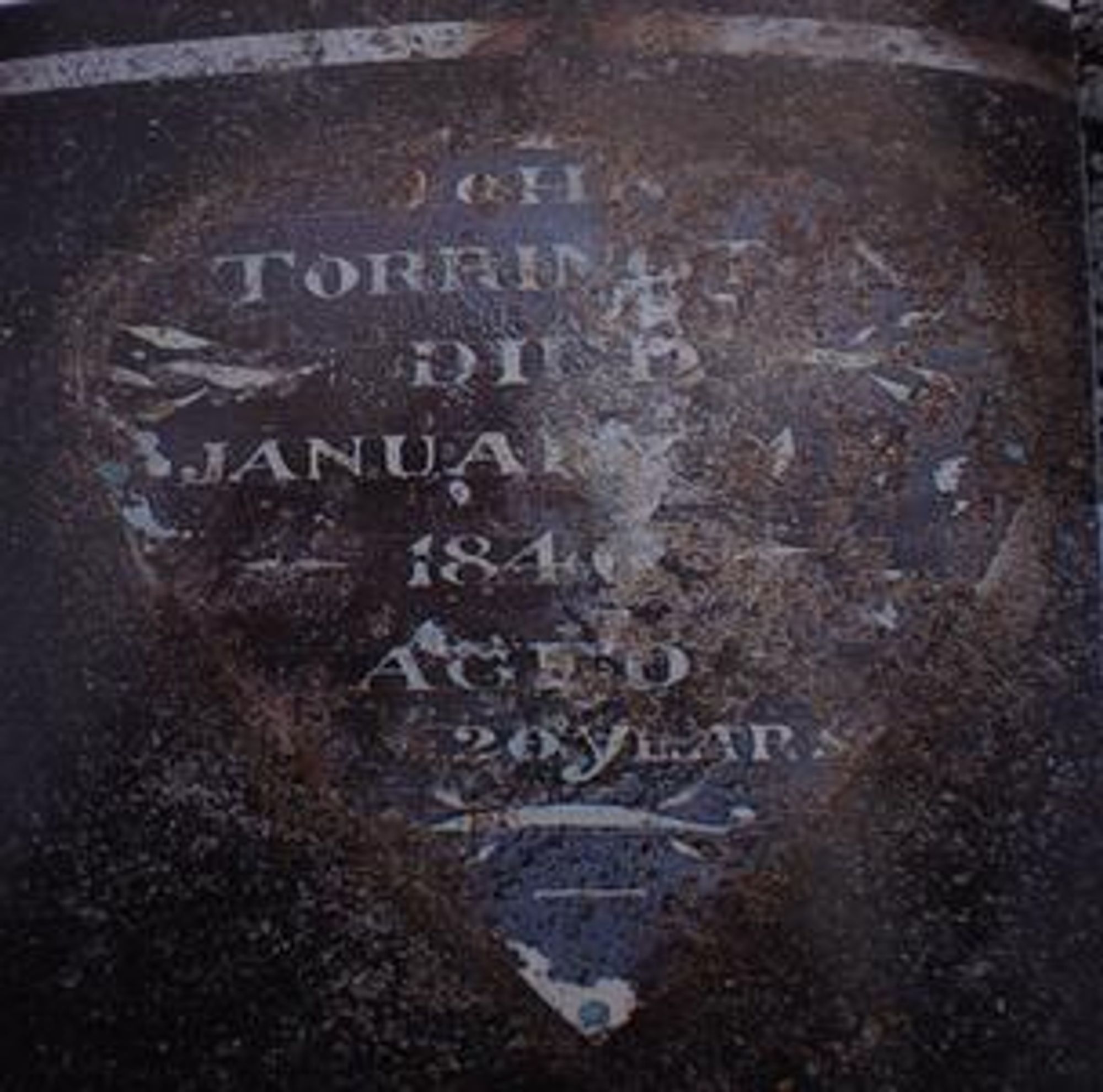 A rusted metal plaque. It's blue-green and has white writing. The writing says, "JOHN TORRINGTON DIED JANUARY 1ST 1846 AGED 20 YEARS."