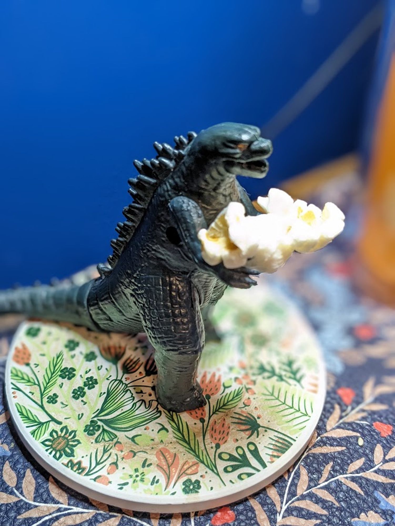 Desk Godzilla, a ferocious green plastic toy about two inches tall, stands on a floral green and pink coaster, cradling a single popped kernel of popcorn in his greedy little arms