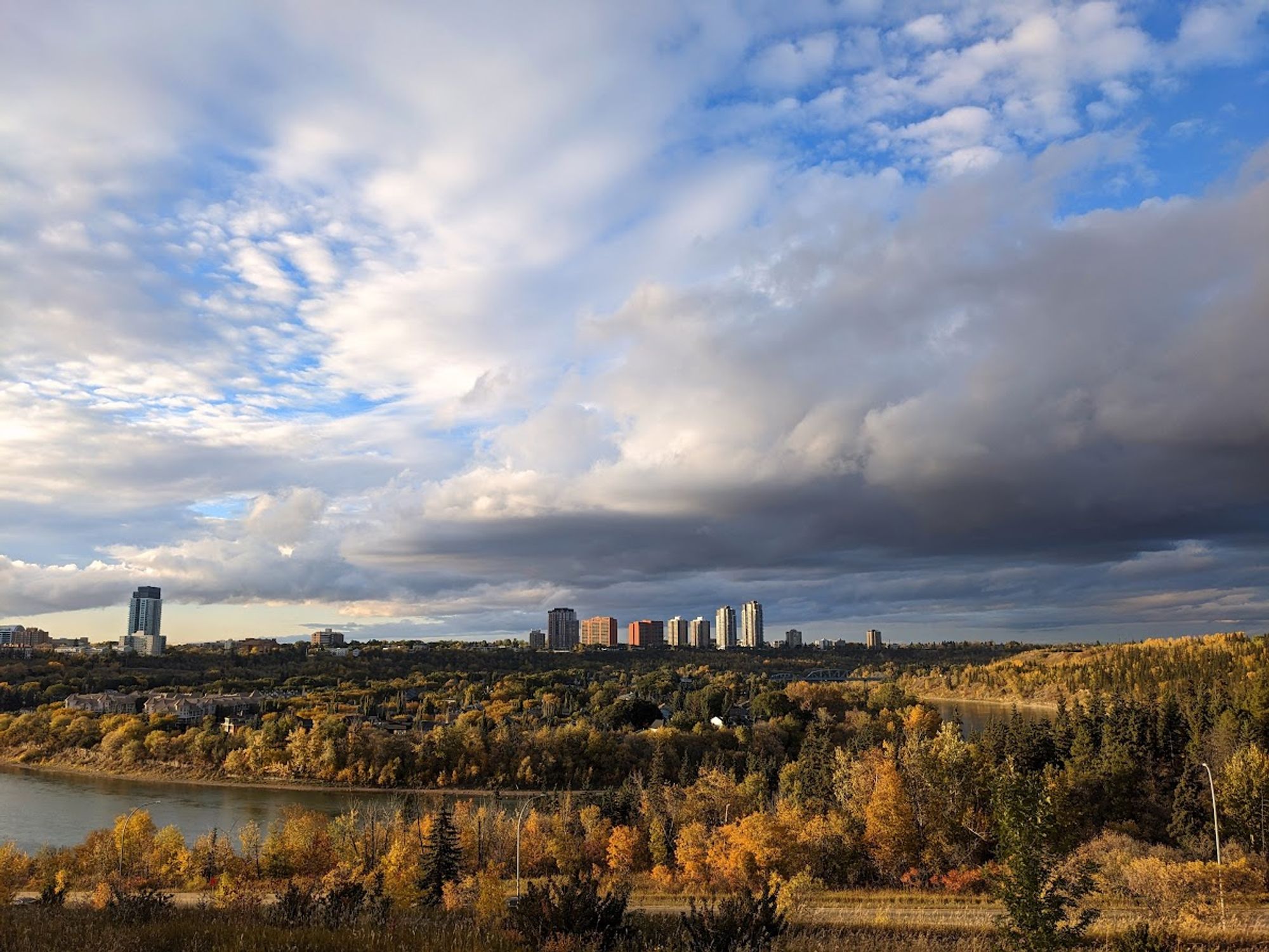 part of edmonton's river valley, showing part of the skyline, a lot of sky with some dark clouds and blue sky, and trees on either side of the bank changing colour for fall