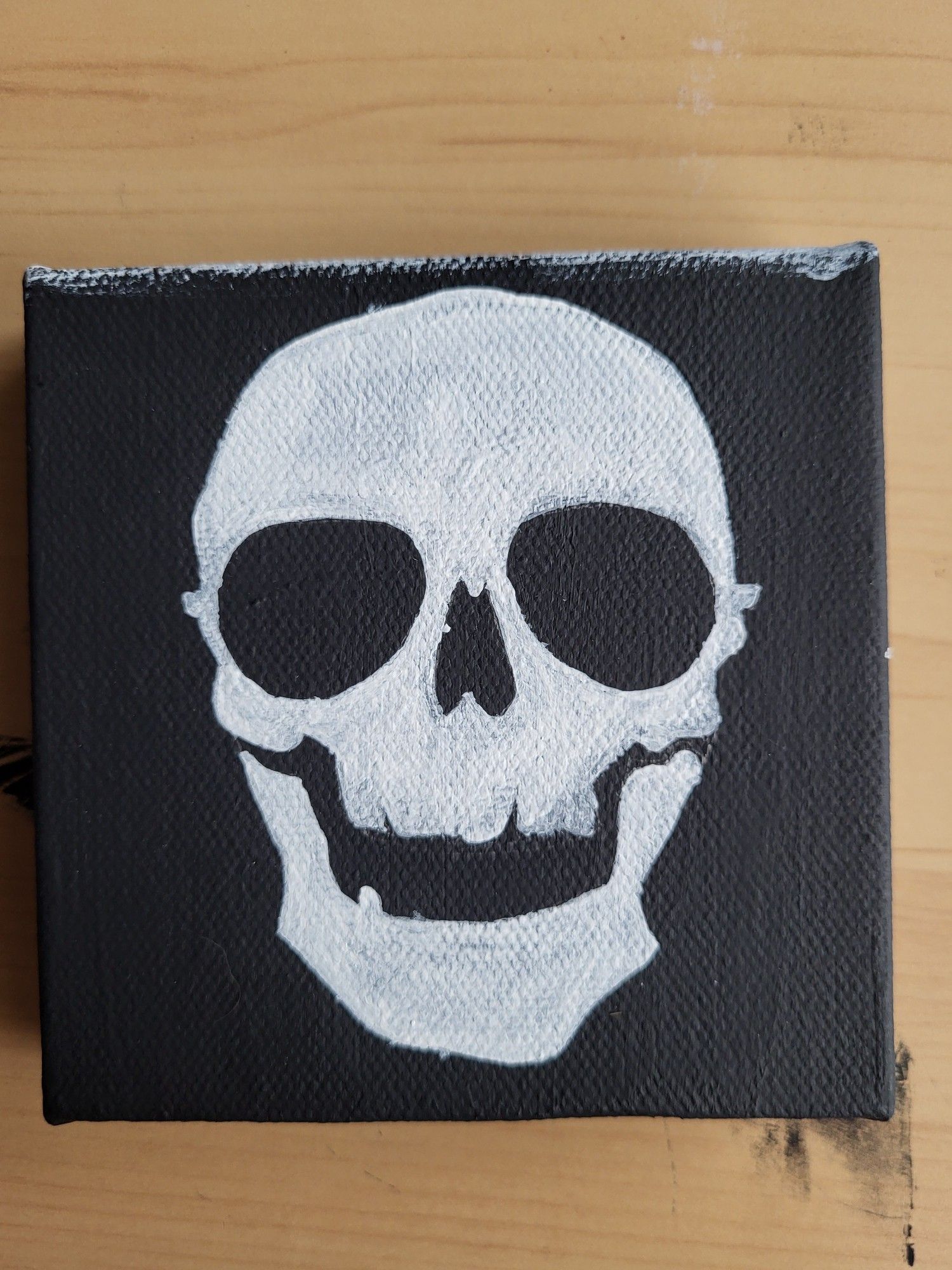 a small black canvas with a white skull painted on a bit loosely. it has its mouth slightly ajar and the eye sockets are too big. it's a rough version of Gideon's skull from The Locked Tomb series