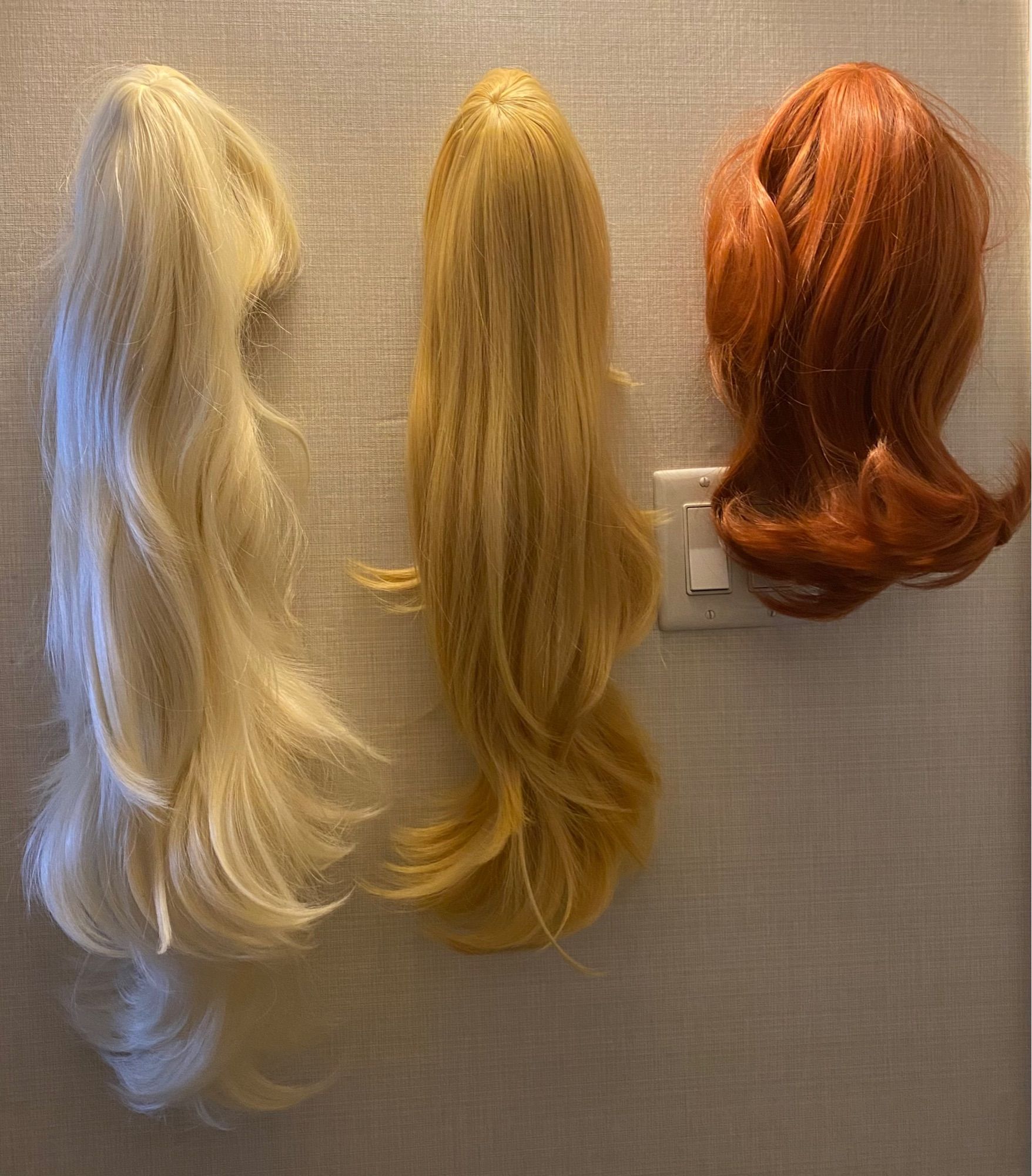 A light blonde wig, a yellow blonde wig, and a ginger wig hang on a wall