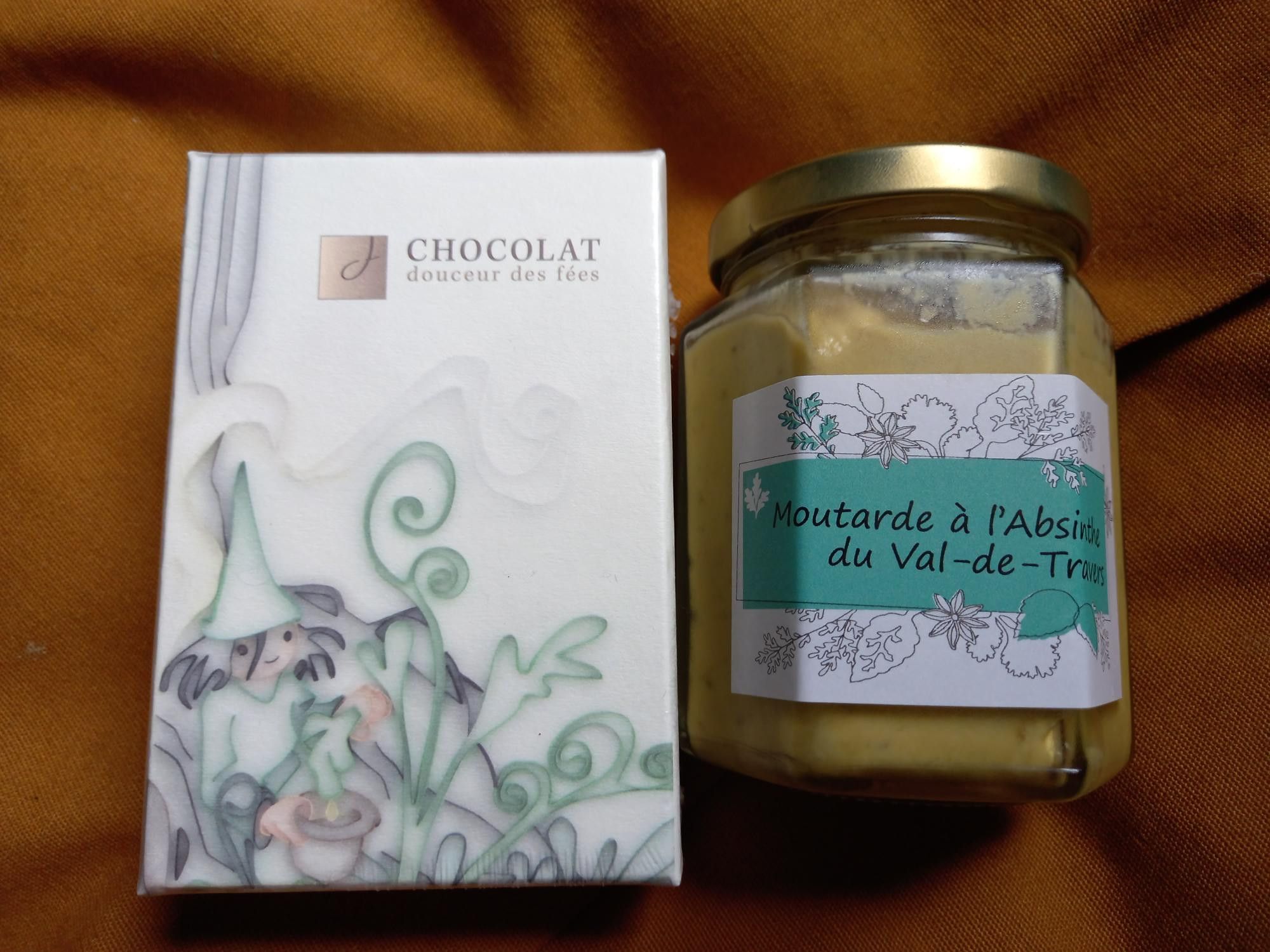 The picture shows two products on a dark yellow background. On the left is a small white box of chocolates - "Chocolat, douceur des fées", with a green fairy on it. On the right is a glass, with a label that says: Moutarde à l'Absinthe du Val-de-Travers".