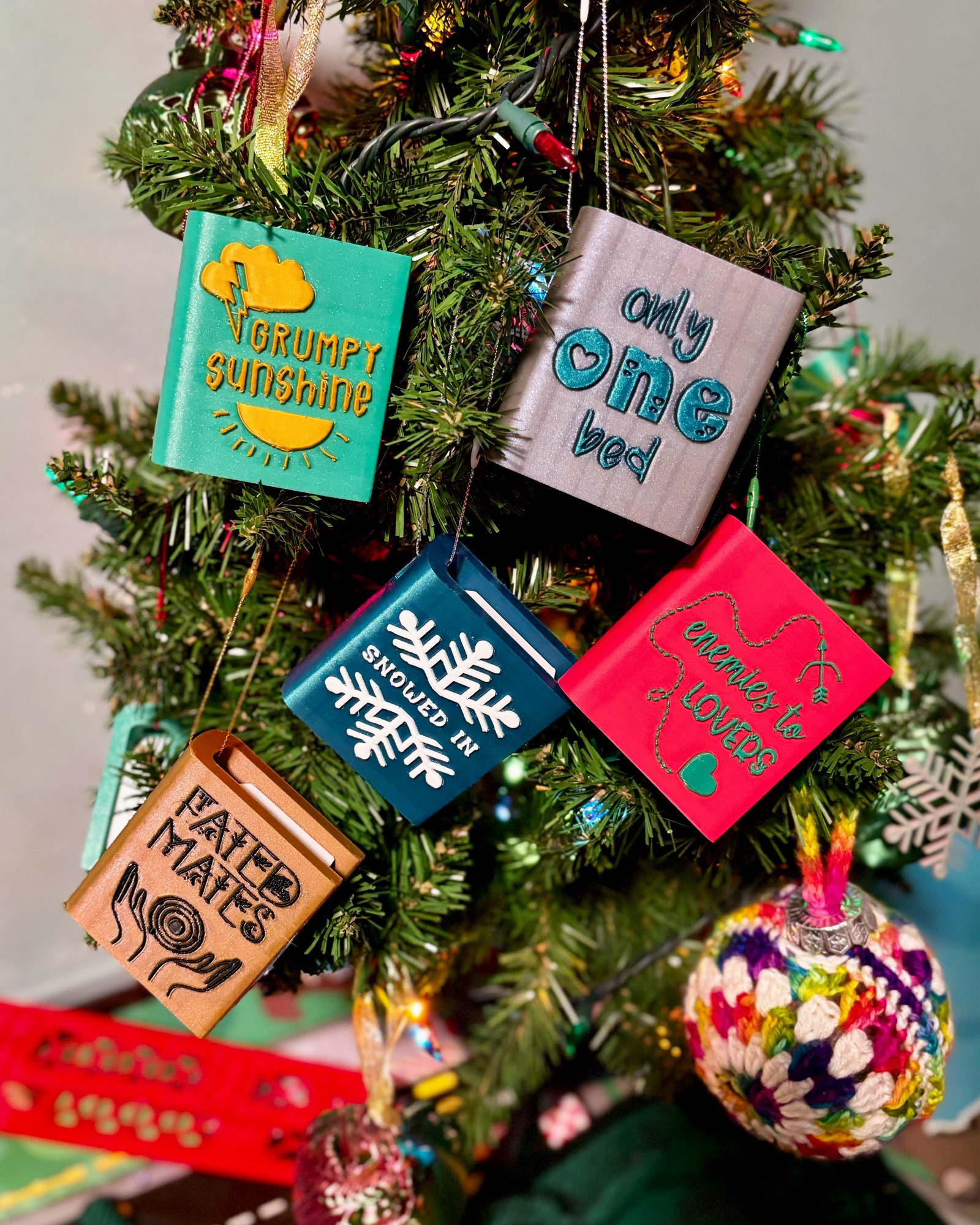 Several book shaped ornaments showing romance tropes on the front; they say “grumpy sunshine”, “only one bed”, “snowed in”, “enemies to lovers”, and “fated mates”