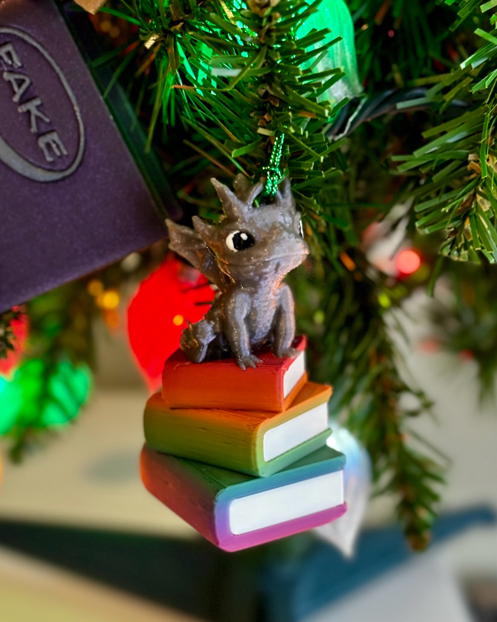 A small 3d printed Christmas ornament that is a silver dragon sitting on a stack of rainbow books 