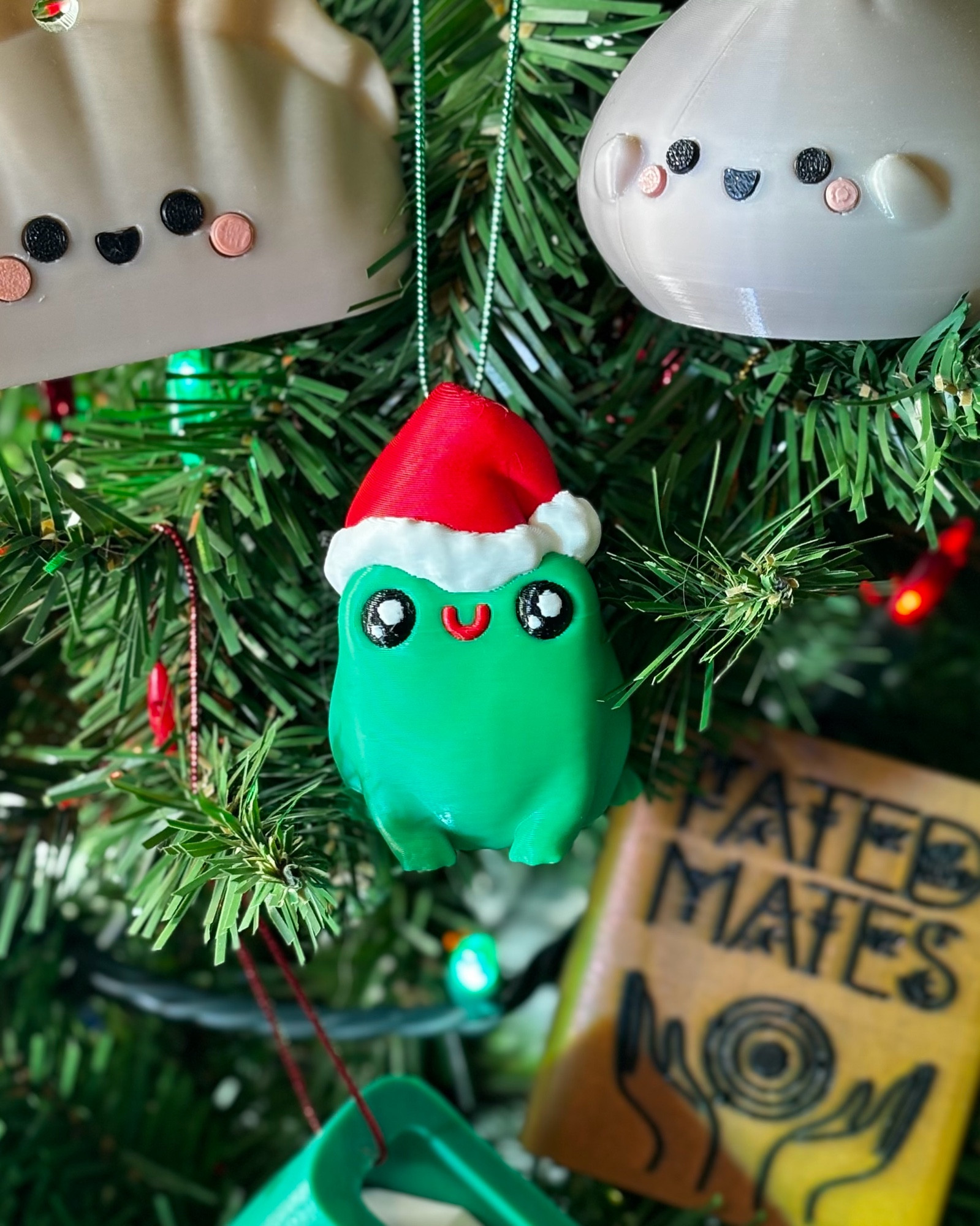 A small 3d printed green frog wearing a Santa hat, hung on a Christmas tree