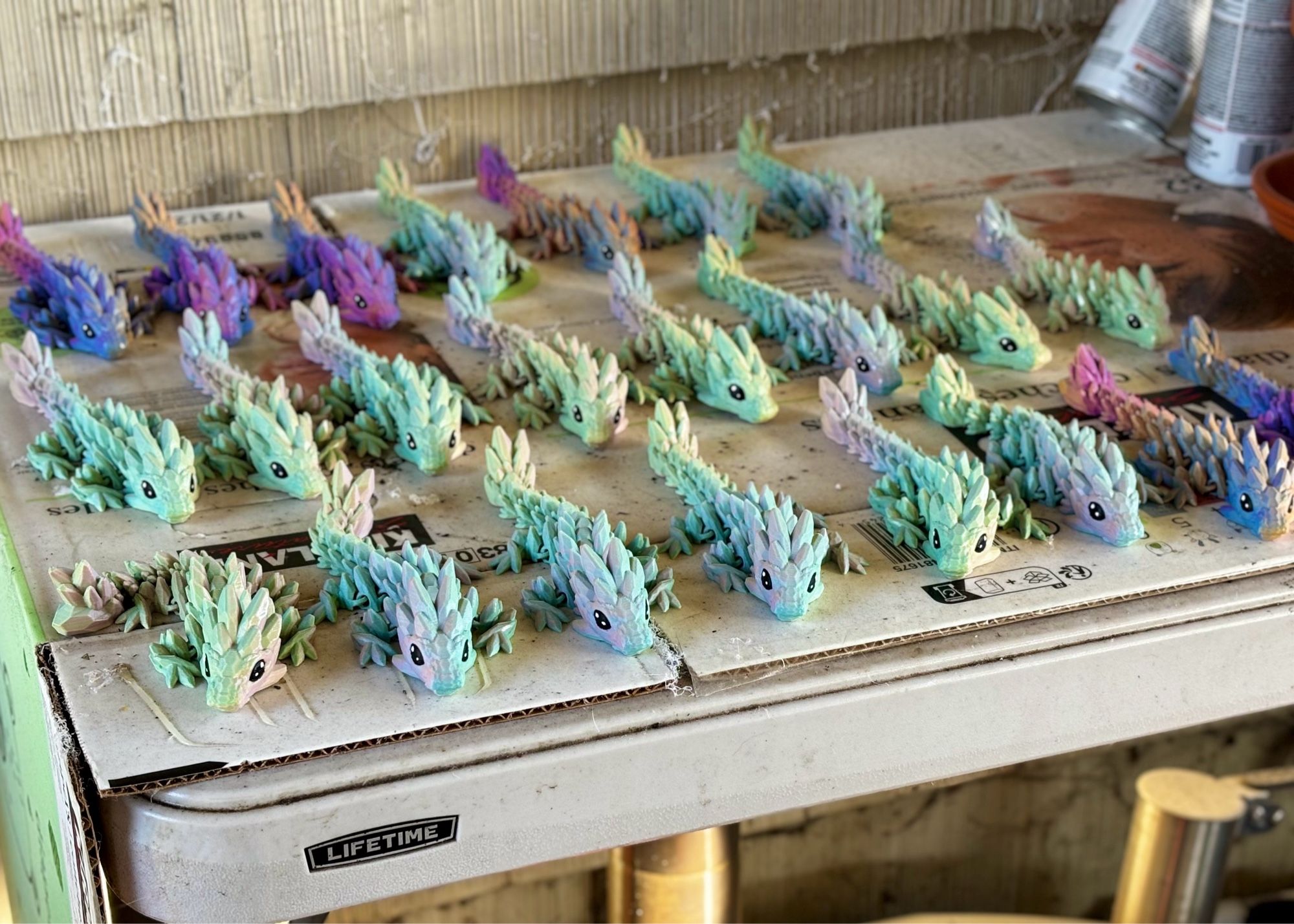 Three rows of several 3d printed dragons printed in either mint or purple filament. They are lined up on a table lined with cardboard