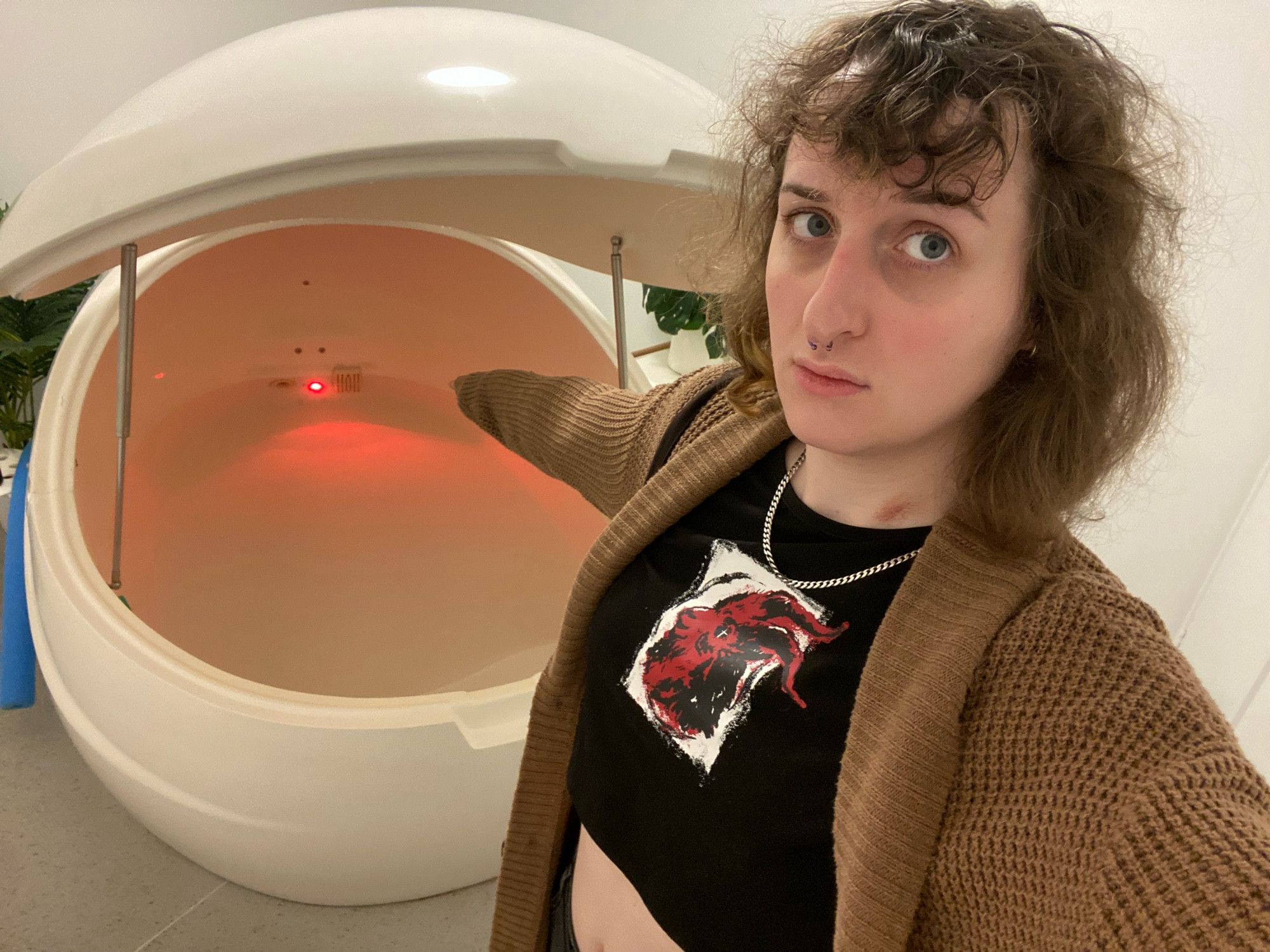 cassie points at a salt float pod with a red light. she is wearing a brown cardigan and black crop top, and looks concerned