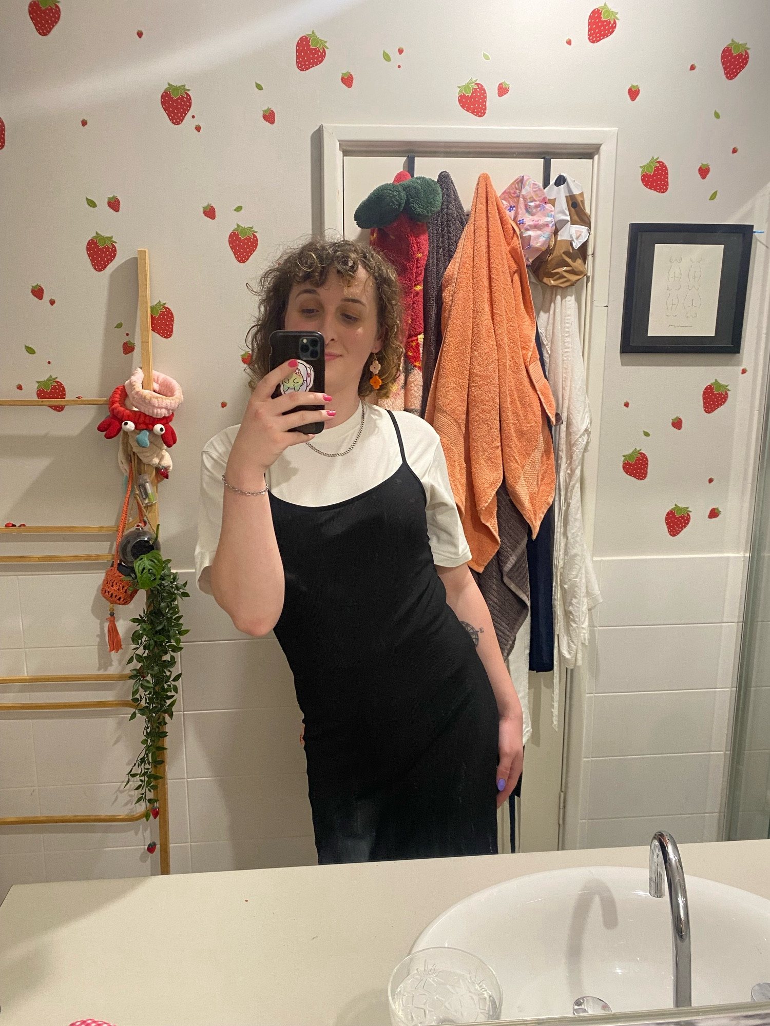 cassie takes a selfie in a bathroom decorated with strawberries on the walls. she’s wearing a white shirt under a black dress, with a chain hanging from her neck, and dangly colourful earrings