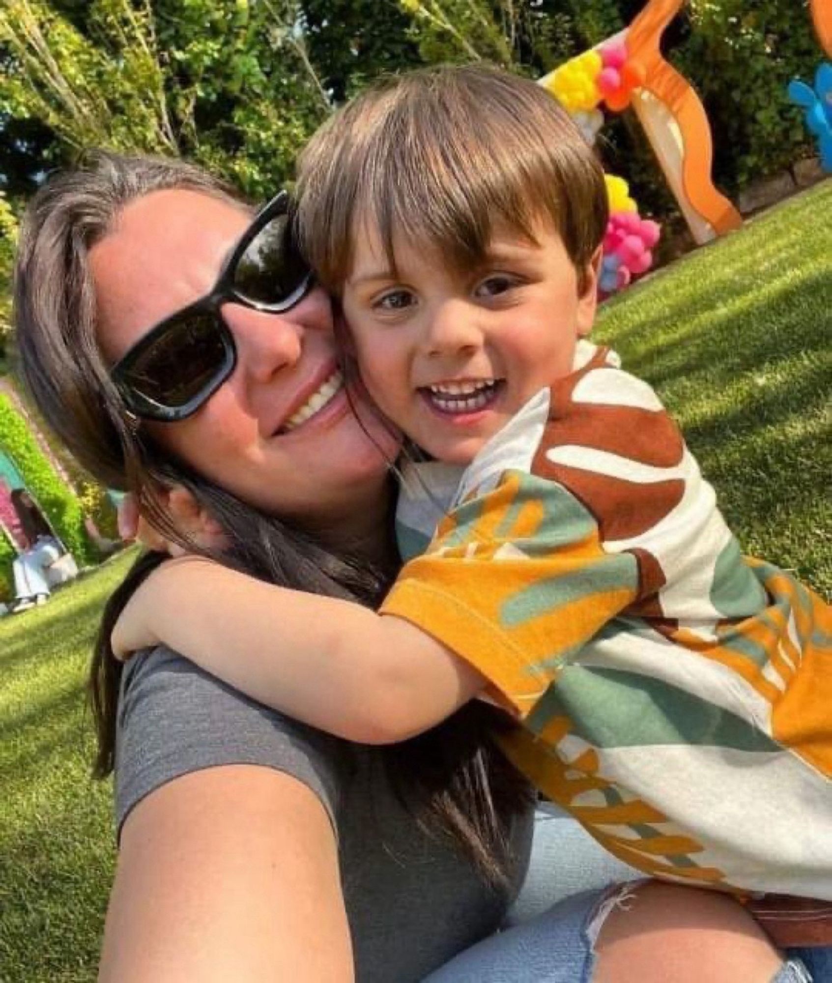 Color photo of a young mother with light skin and light brown hair smiling and holding her young son who is hugging her and smiling broadly at the camera. They are outside with green grass in the background.