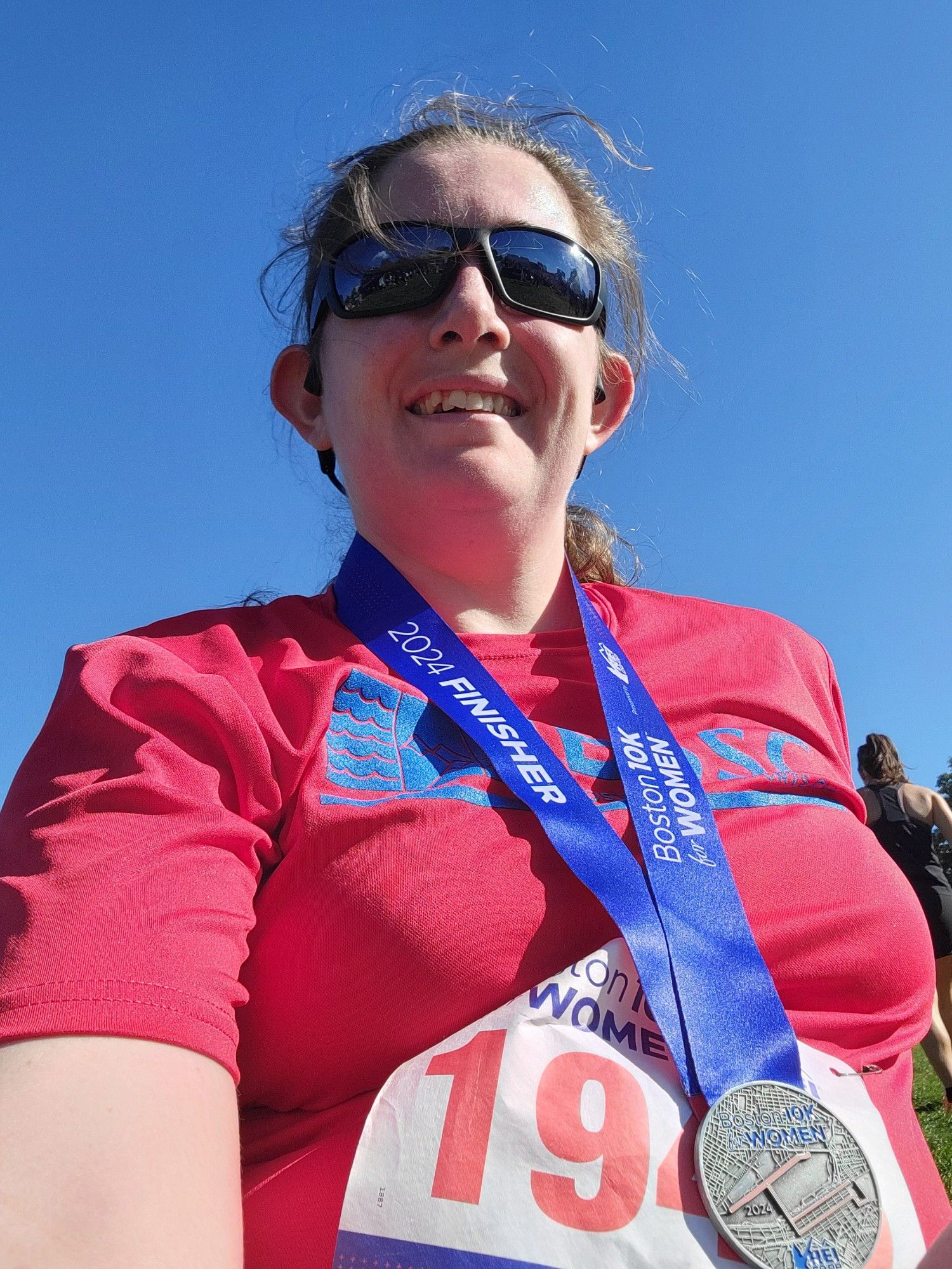 Photo of me, a white woman with her brown hair pulled back into a ponytail, wisps flying all over, wearing sunglasses, a red shirt, and a finishers medal, with the race bib attached to the shirt