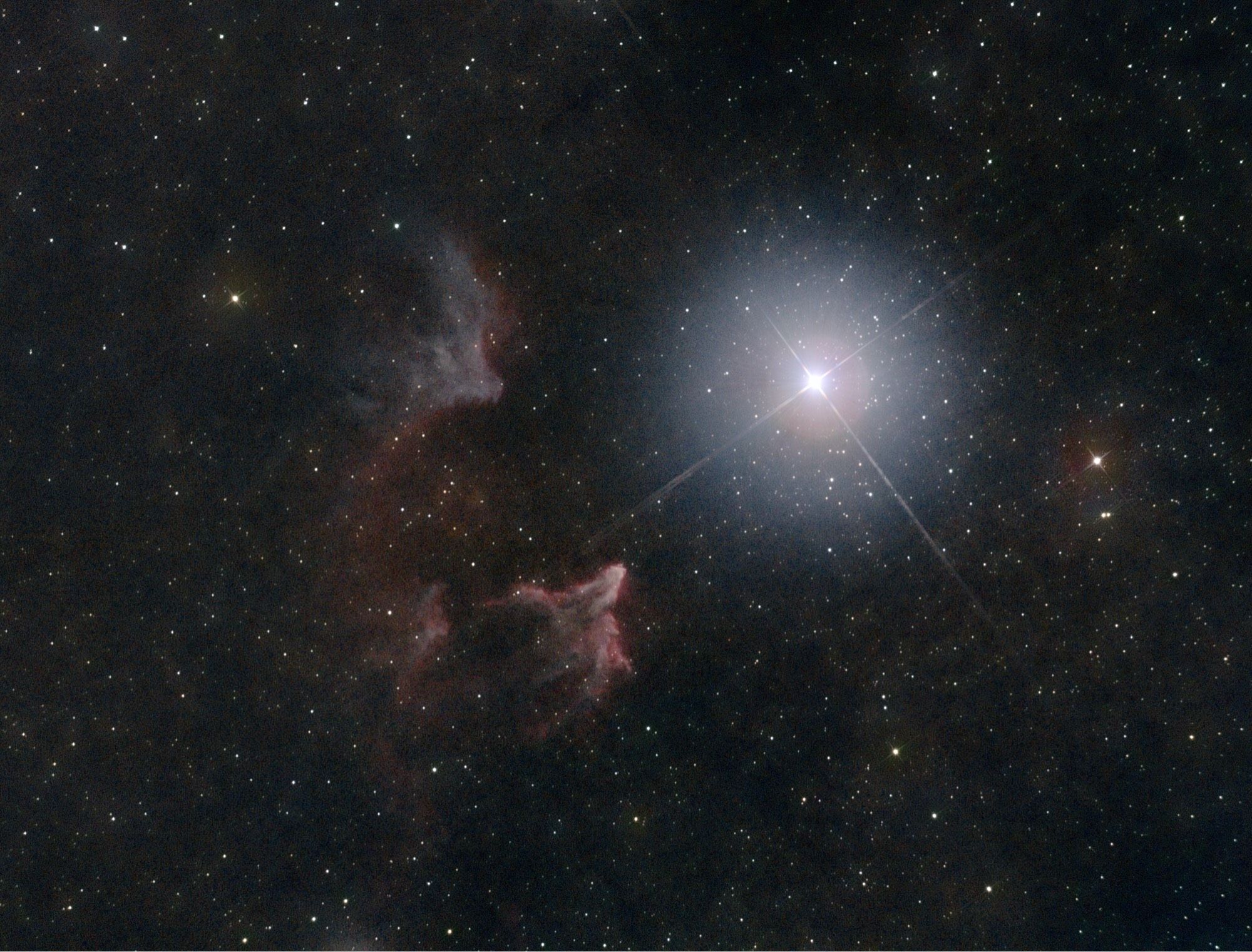 A bright white star with a hazy aura and four long diffraction spikes, to the right of a ghostly pair of nebulae, one mostly greyish, the other, below it, tinged with pinks and reds, all against a dark background sprinkled with little stars.