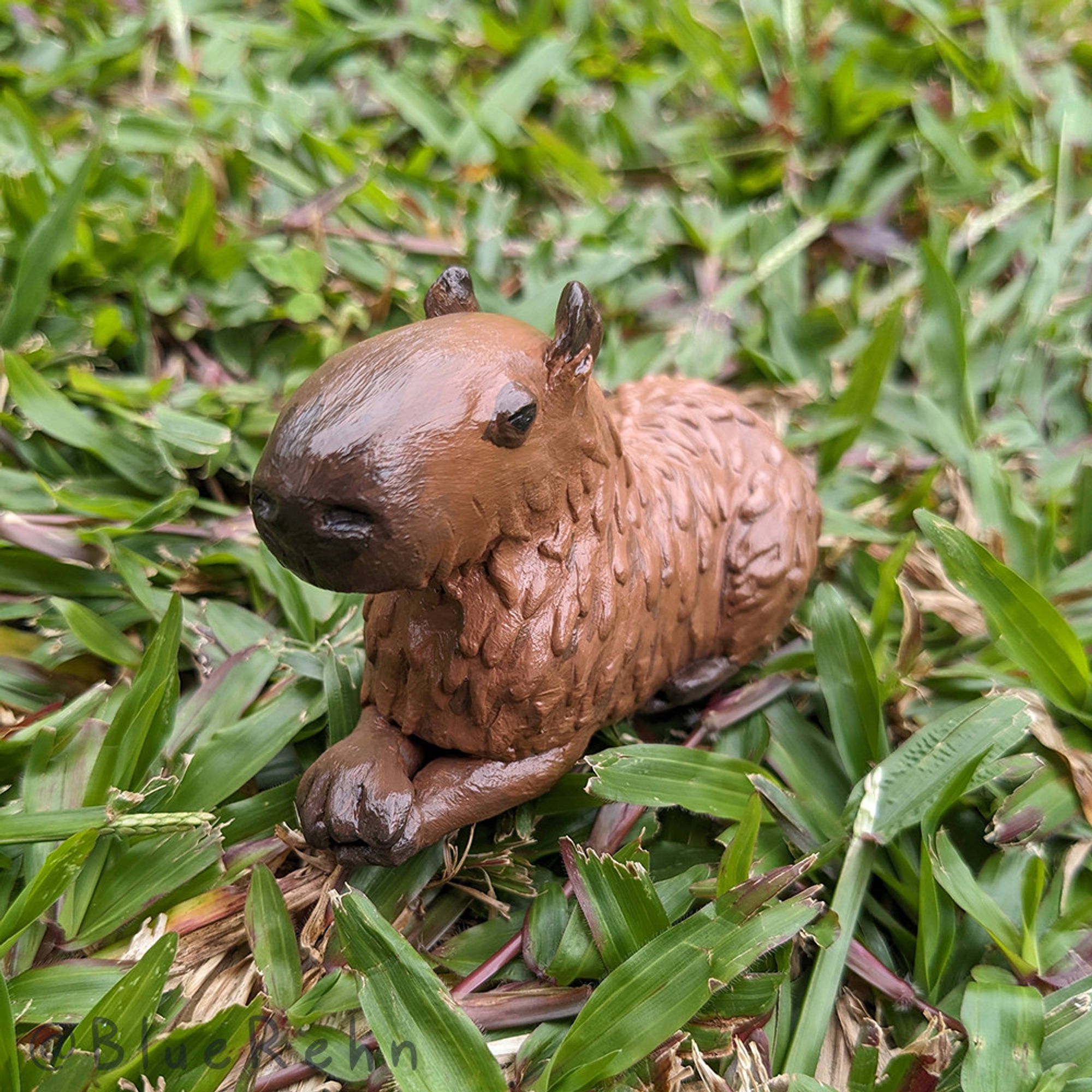 A photograph of a small clay capybara resting on grass, facing towards the camera