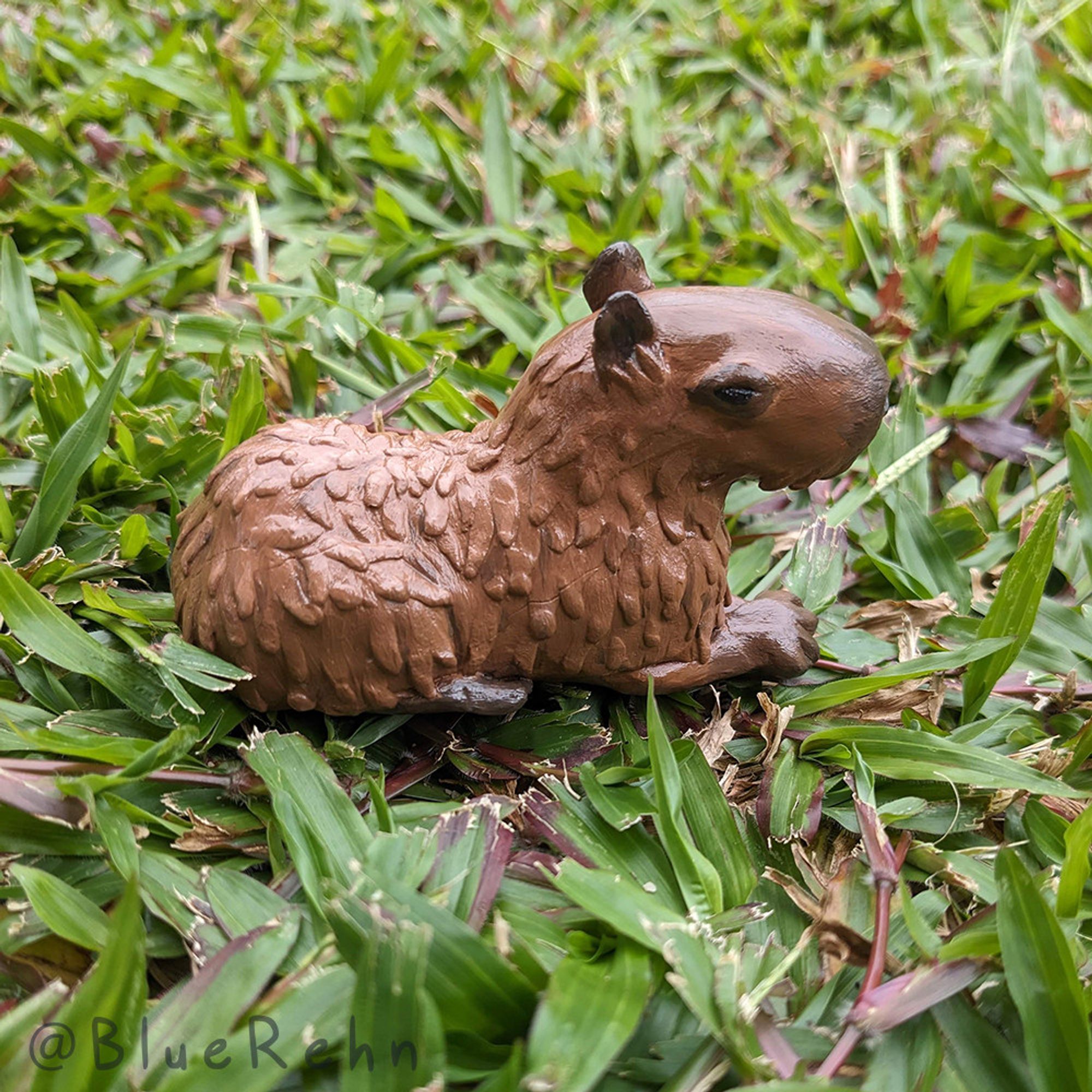 A photograph of a small clay capybara resting on grass