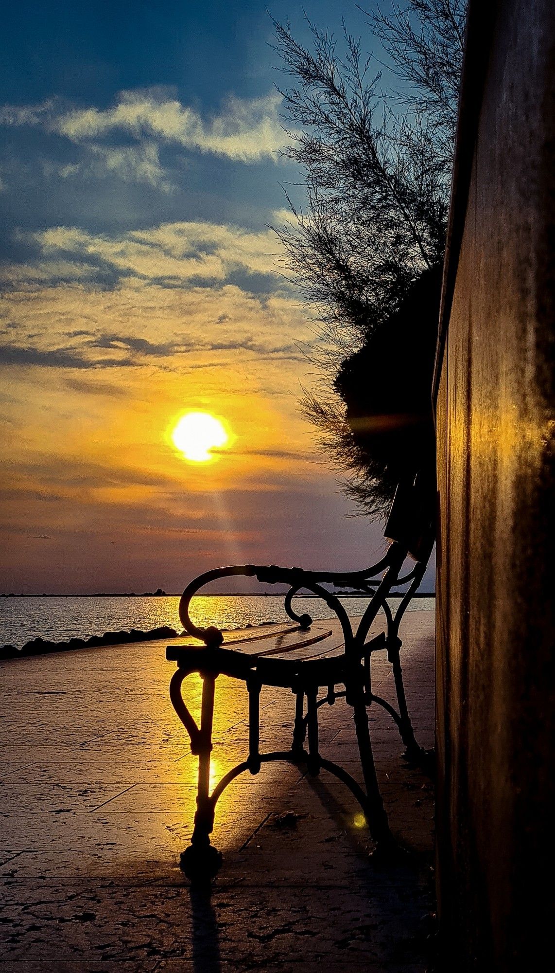 Eine Bank steht auf einer Promenade am Meer.  Im Sonnenuntergang spiegelt sich die Sonne auf dem Wasser und dem Sternen.