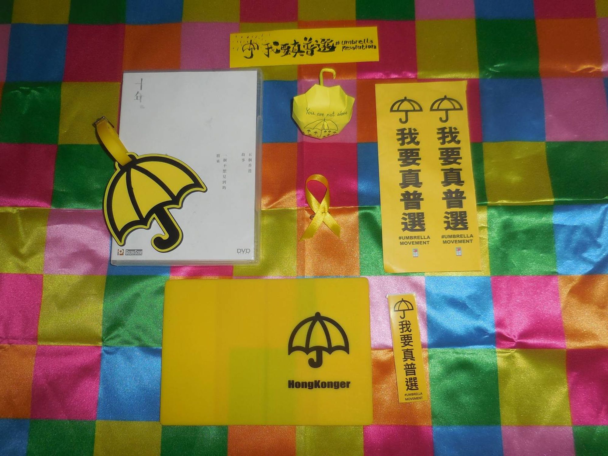 Various Umbrella Movement & Umbrella Revolution stickers, a yellow ribbon, an umbrella suitcase tag on a ten years dvd, a Hongkonger passport cover and a litte yellow paper umbrella with umbrellas painted on it and the slogan You are not alone on a Lennon Wall flag.