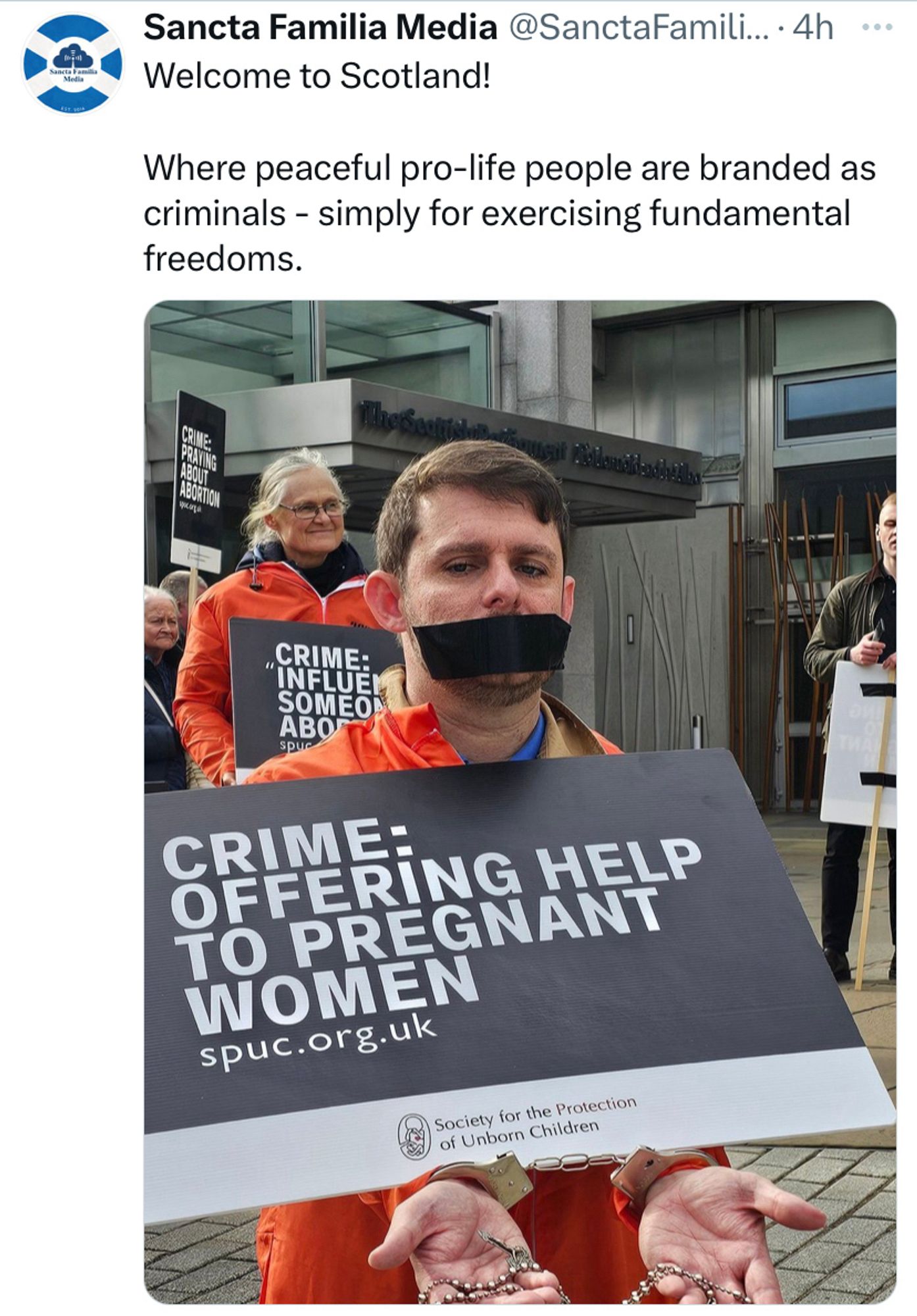 Man in orange jumpsuit with handcuffs and gag he put on himself, holding a sign reading. ‘Crime: offering help to pregnant women’