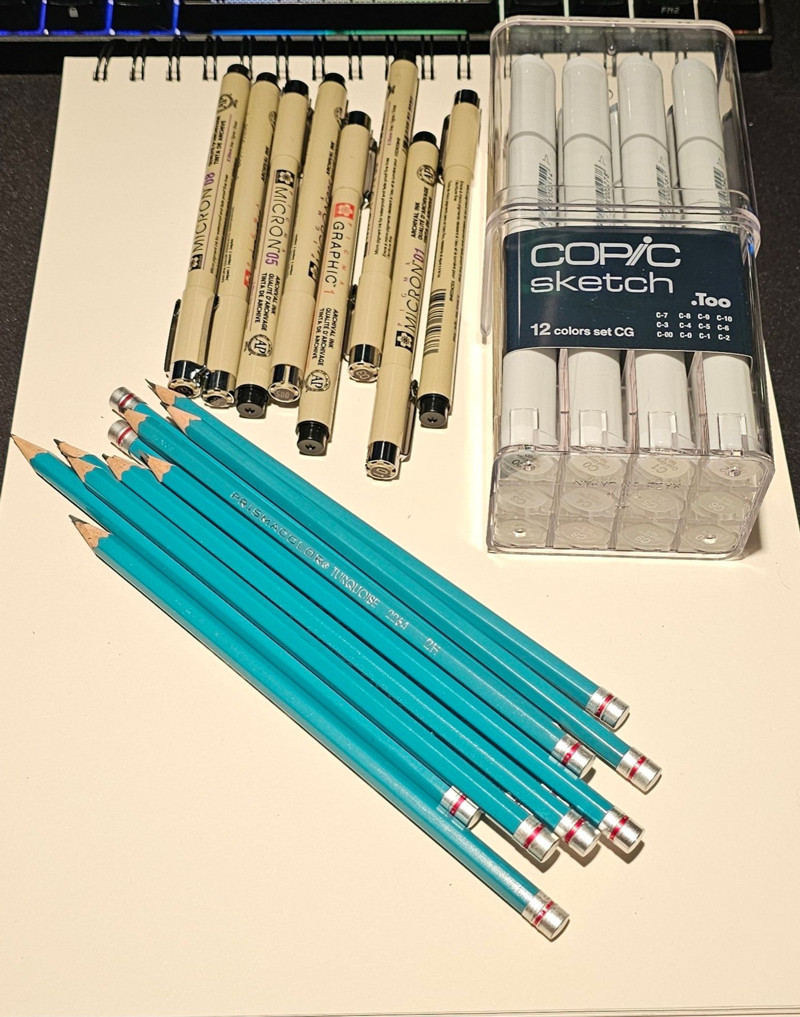 An image of a drawing pad. On top of the drawing pad are teal prisma color pencils, drawing pens and a set of cool gray copic markers.