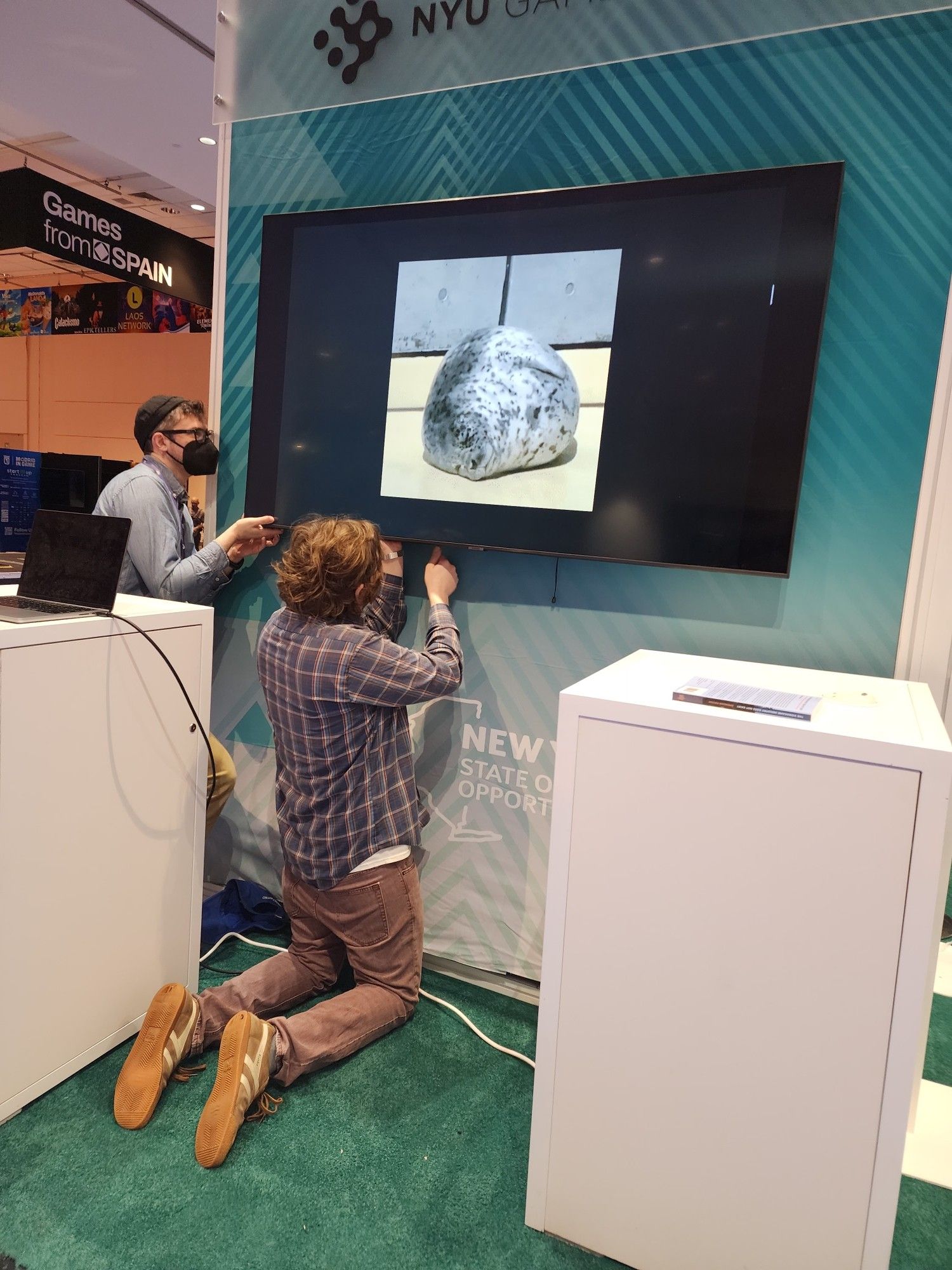 Two men adjusting a monitor for the NYU Game Center's Game Developers Conference expo booth. Monitor has an image of a chonky seal in full screen.