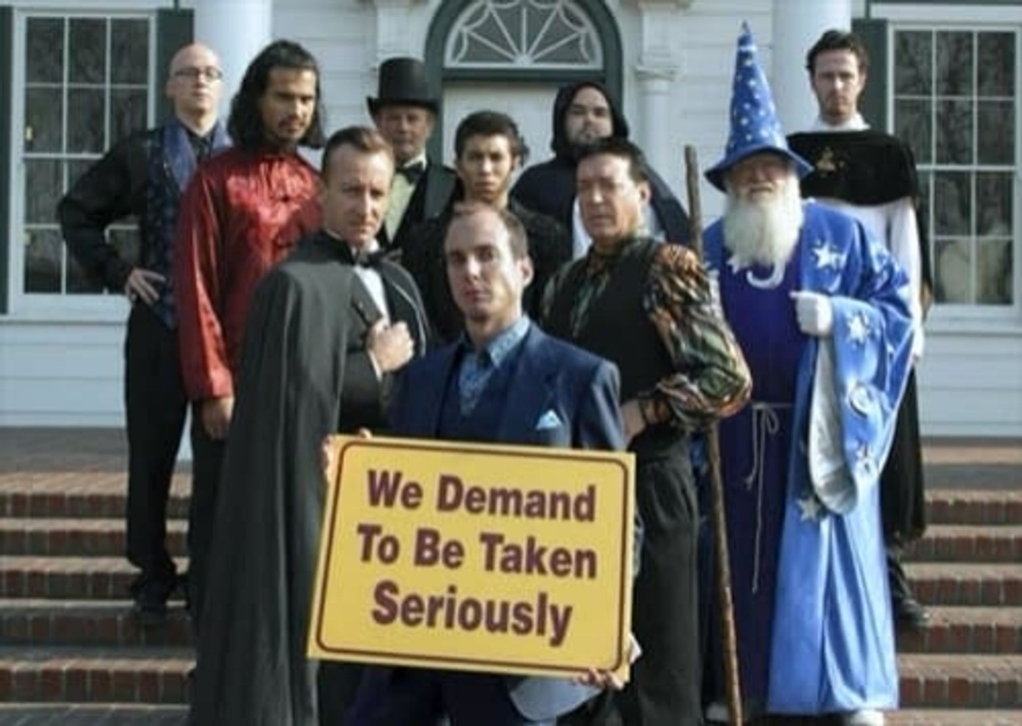 Imagem retirada do seriado Arrested Develpment. Várias pessoas vestidas com tragédia de magos e feiticeiros fazem pose, na frente um homem segura um cartaz escrito "We demand to be taken seriously"