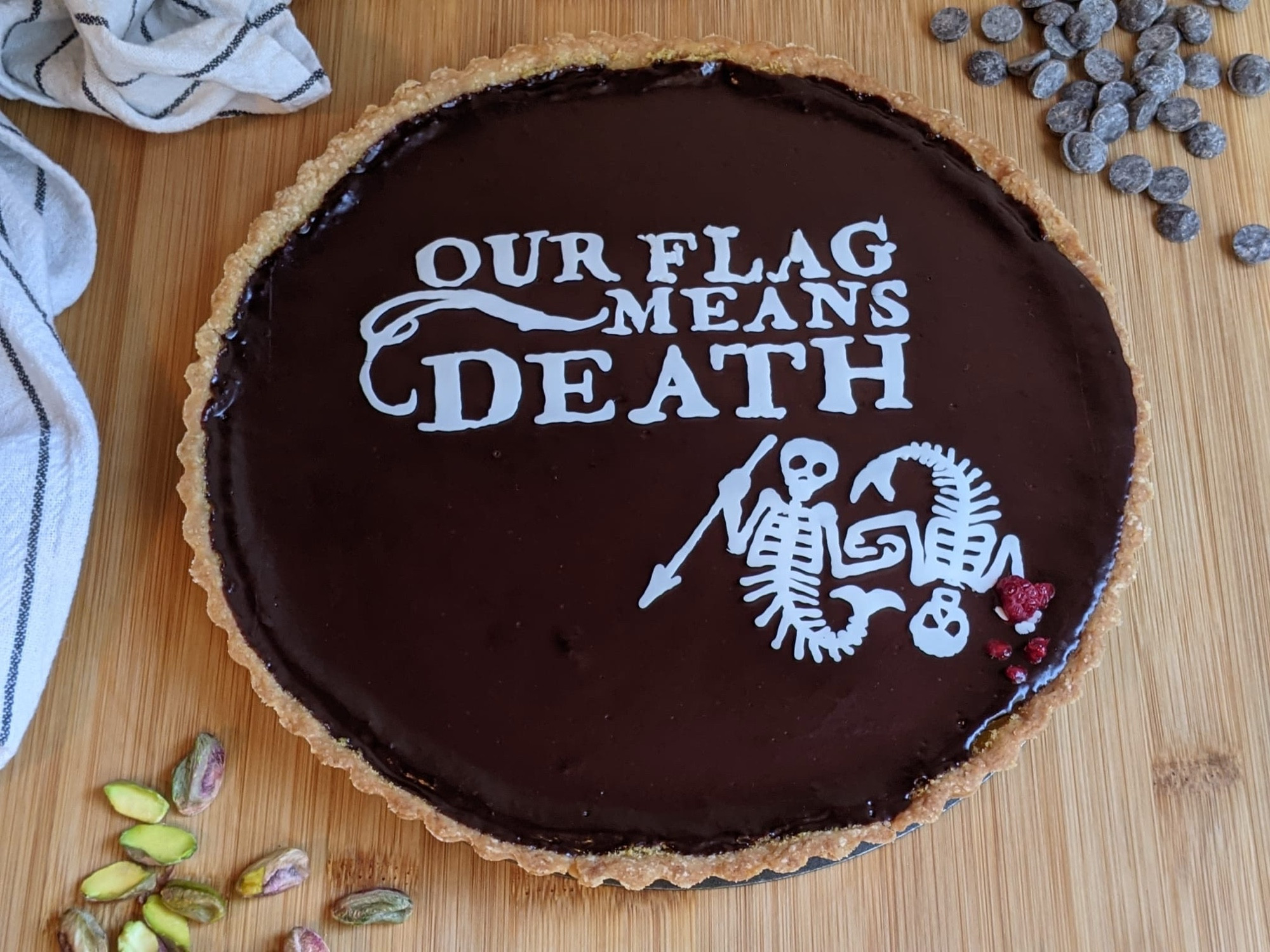 Food, a photo of a chocolate pie with a white chocolate decoration of the "our flag means death" logo and two skeleton mermaids