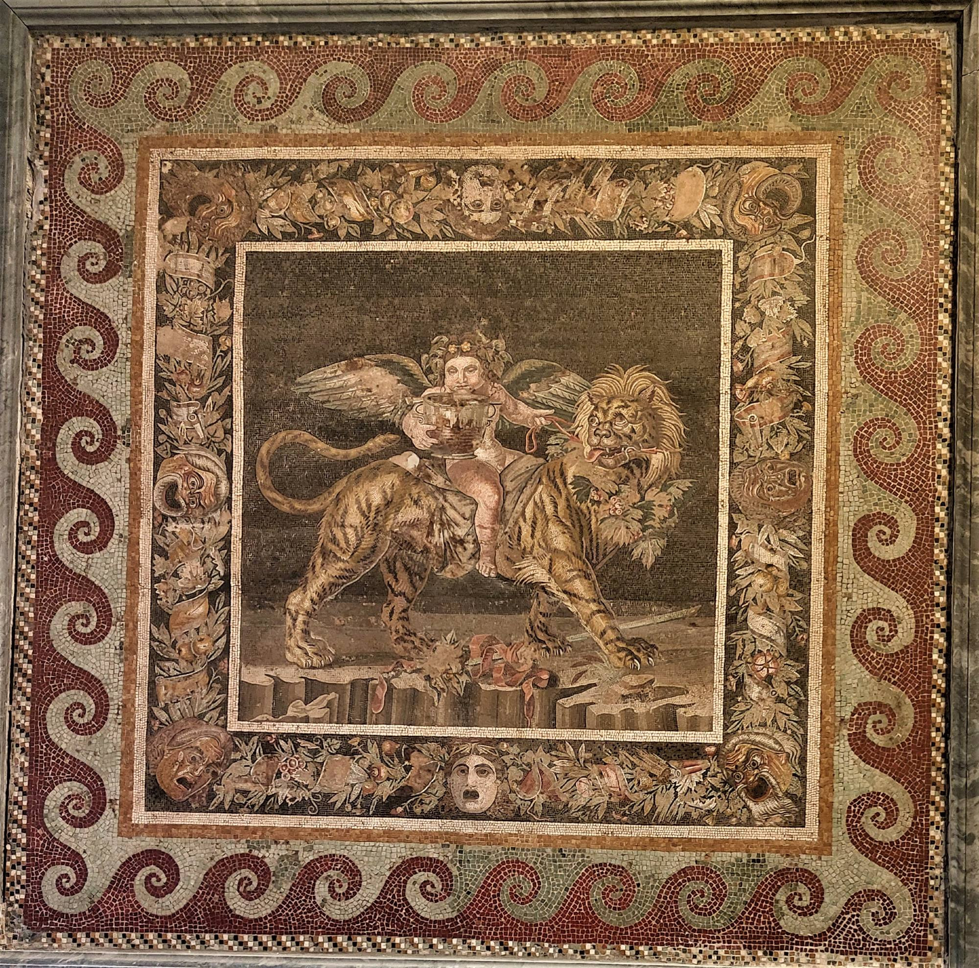 A scene of a young winged god - probably Dionysus - on the back of a tiger with a lion's head. Gripping the harness of the beast, he wears a garland of fruit and leaves on his head and  holds a large basin of wine.
This central image is bounded by an elaborate series of garlands and dramatic masks, all entertwined. 
Outside of this is a wavy border in dark red and off-white. 