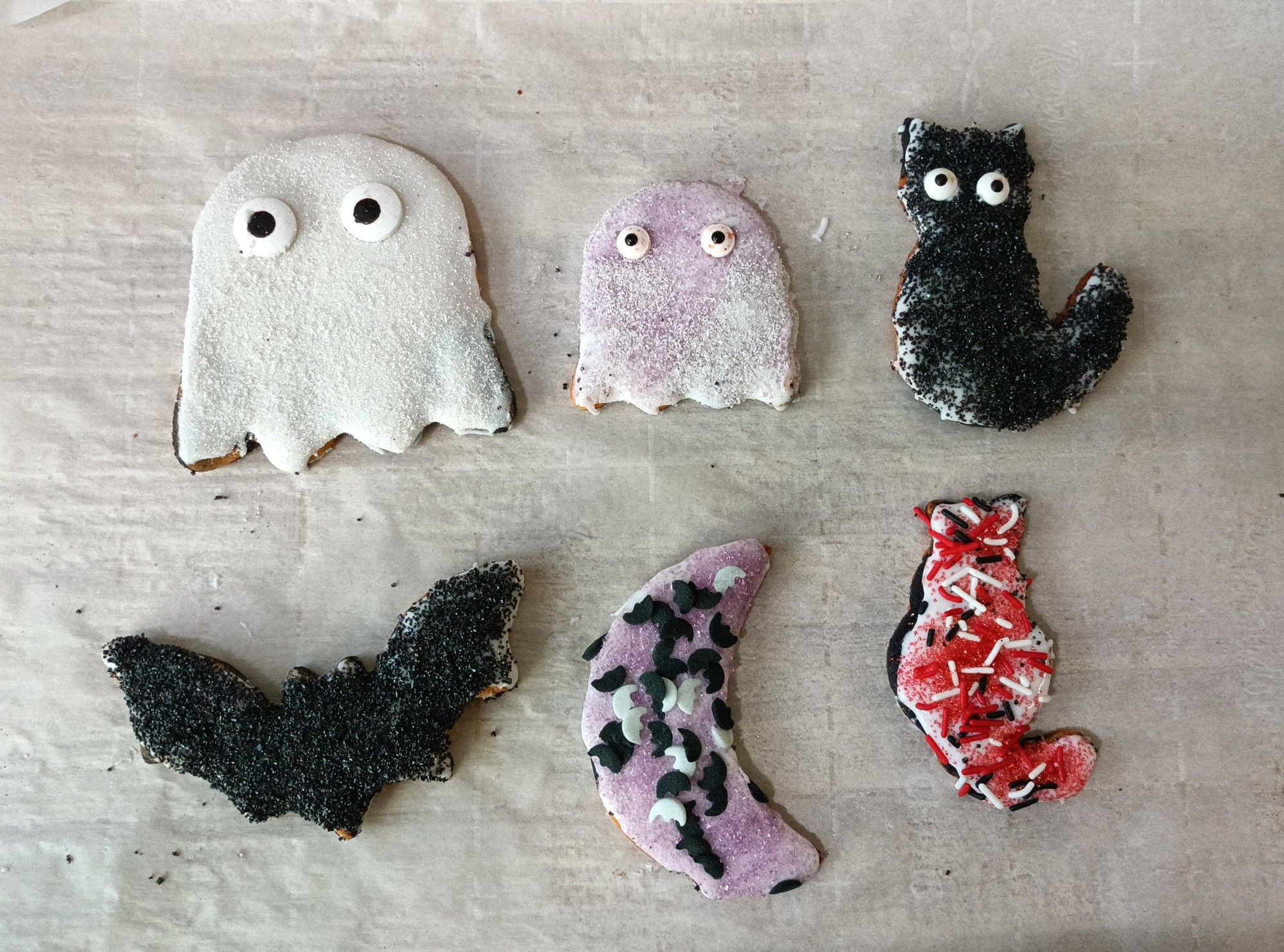 Large ghost with white sugar icing and sugar eyes, small ghost with white and purple sugar icing with sugar eyes, cat cookie with black sugar icing and sugar eyes, bat cookie with black sugar icing, moon sugar cooking with purple sugar icing and sugar moon sprinkles, cat with red, black and white sprinkles. 