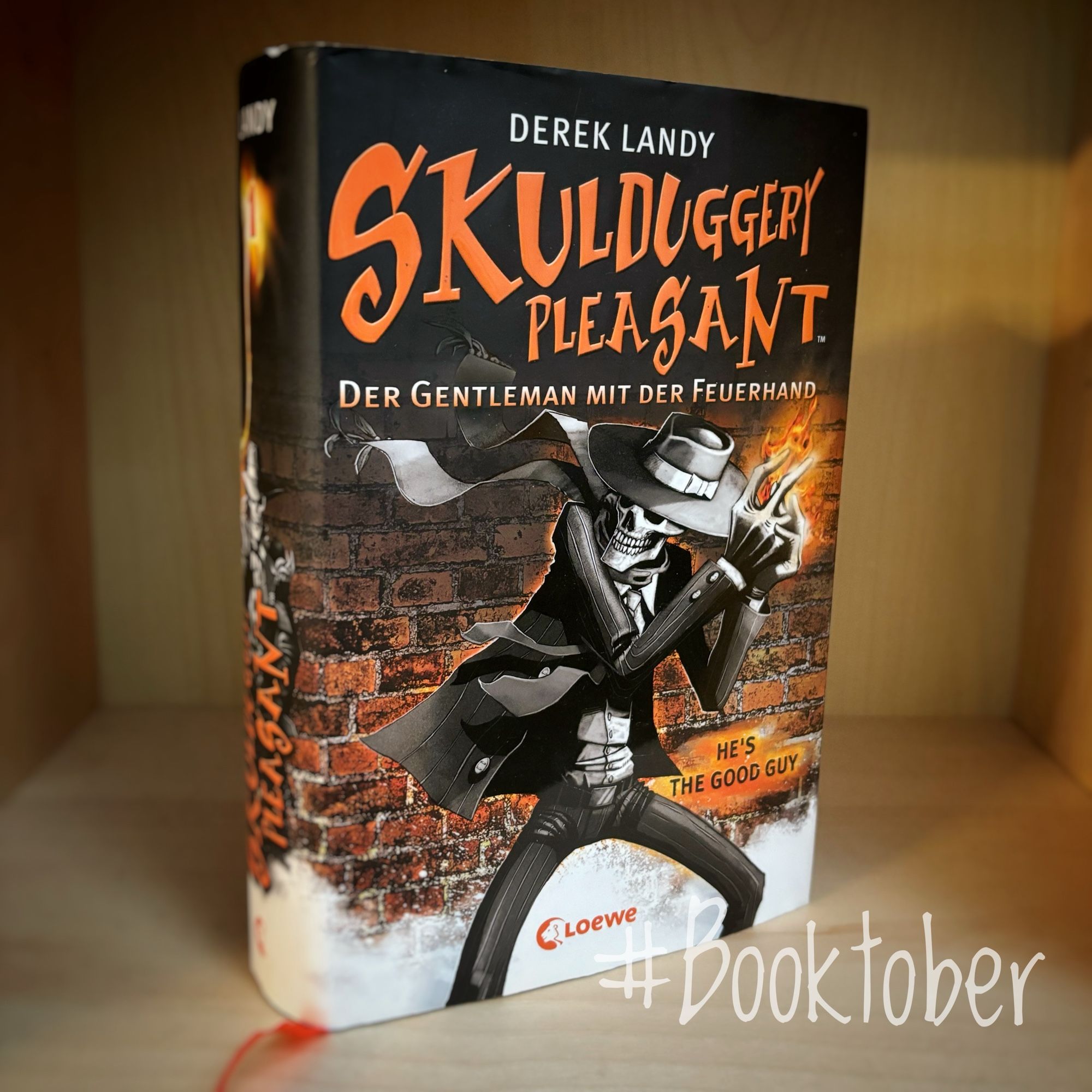 Ein schmales Bücherregalfach in hellem Holz im Hintergrund.
In der Mitte steht das Buch „Skulduggery Pleasant - Der Gentleman mit der Feuerhand“ von Derek Landy aus dem Loewe Verlag.
Im oberen Drittel dominiert in orangefarbener Schrift der Titel plus dem Untertitel darunter. Im unteren Bereich ist besagter Skulduggery Pleasant also Zeichnung abgebildet: Ein Skelett in feinem schwarzen Anzug mit Hut. Aus seinen behandschuhten Händen ragt eine Flamme heraus. Er steht vor einer orange gehaltenen Backsteinwand. 
Unten rechts steht in weißer, leicht durchscheinender Schrift „#Booktober".