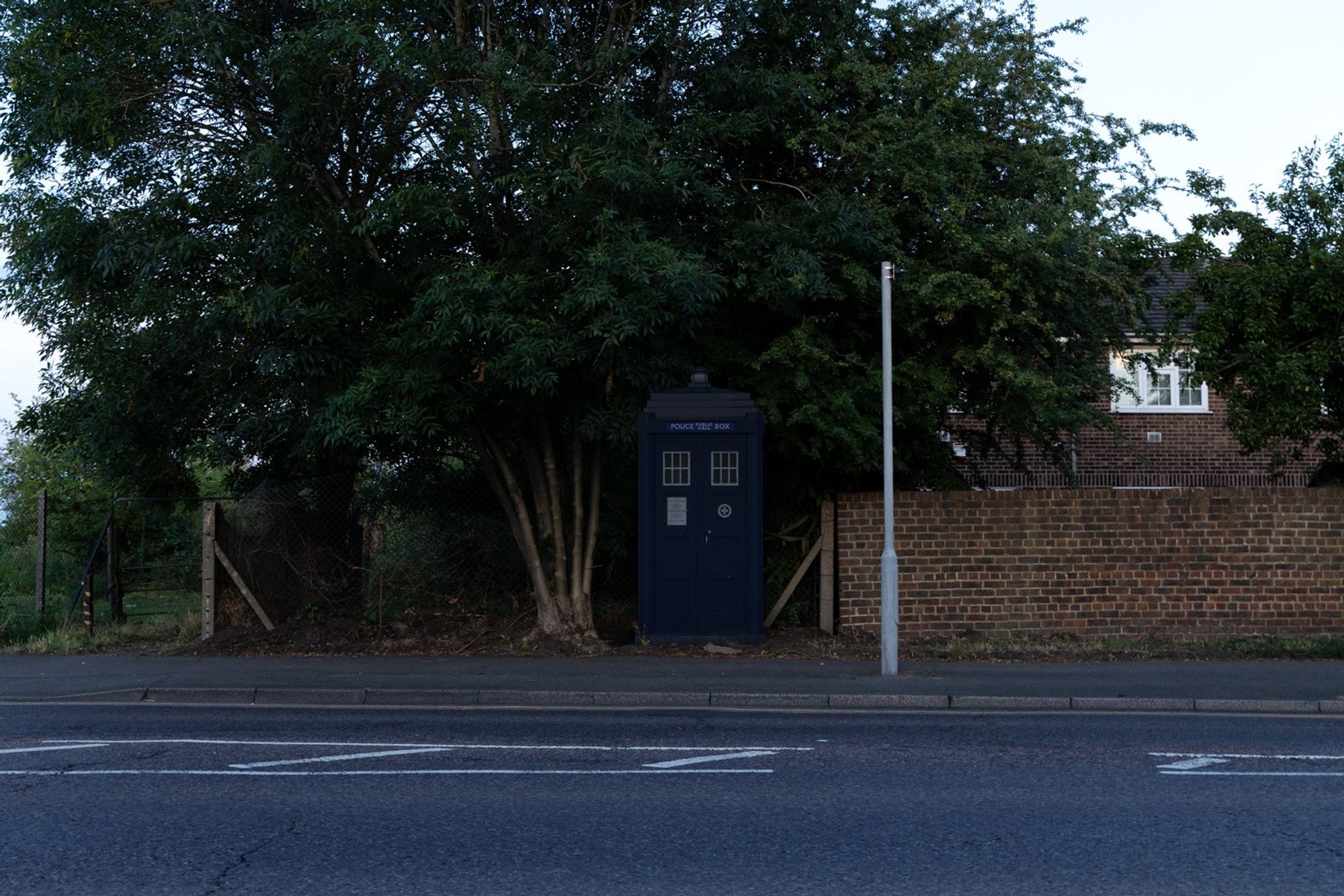 3D composite of a police box under some trees and beside a wall, with a road in the foreground.