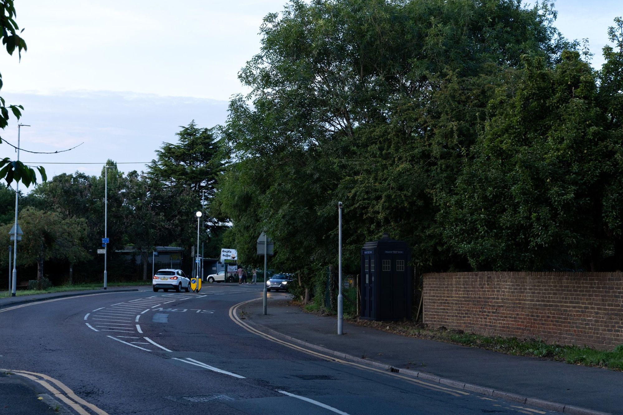 3D composite of a police box under some trees and beside a wall on the right of the photo, with a road in front of it on the left.