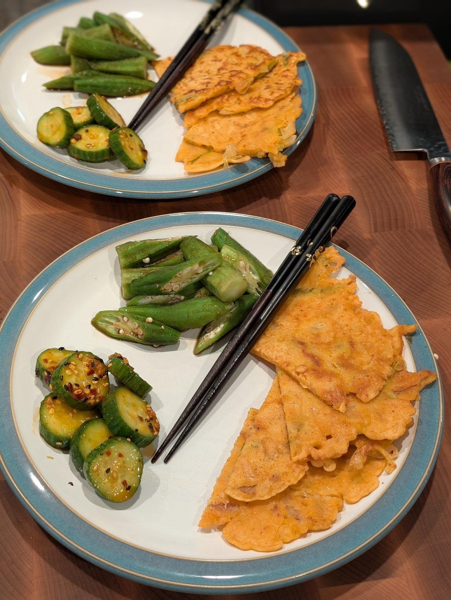 Two dinner plates, each containing slices of a crispy jeon, some spicy marinated cucumber, and dressed okra.