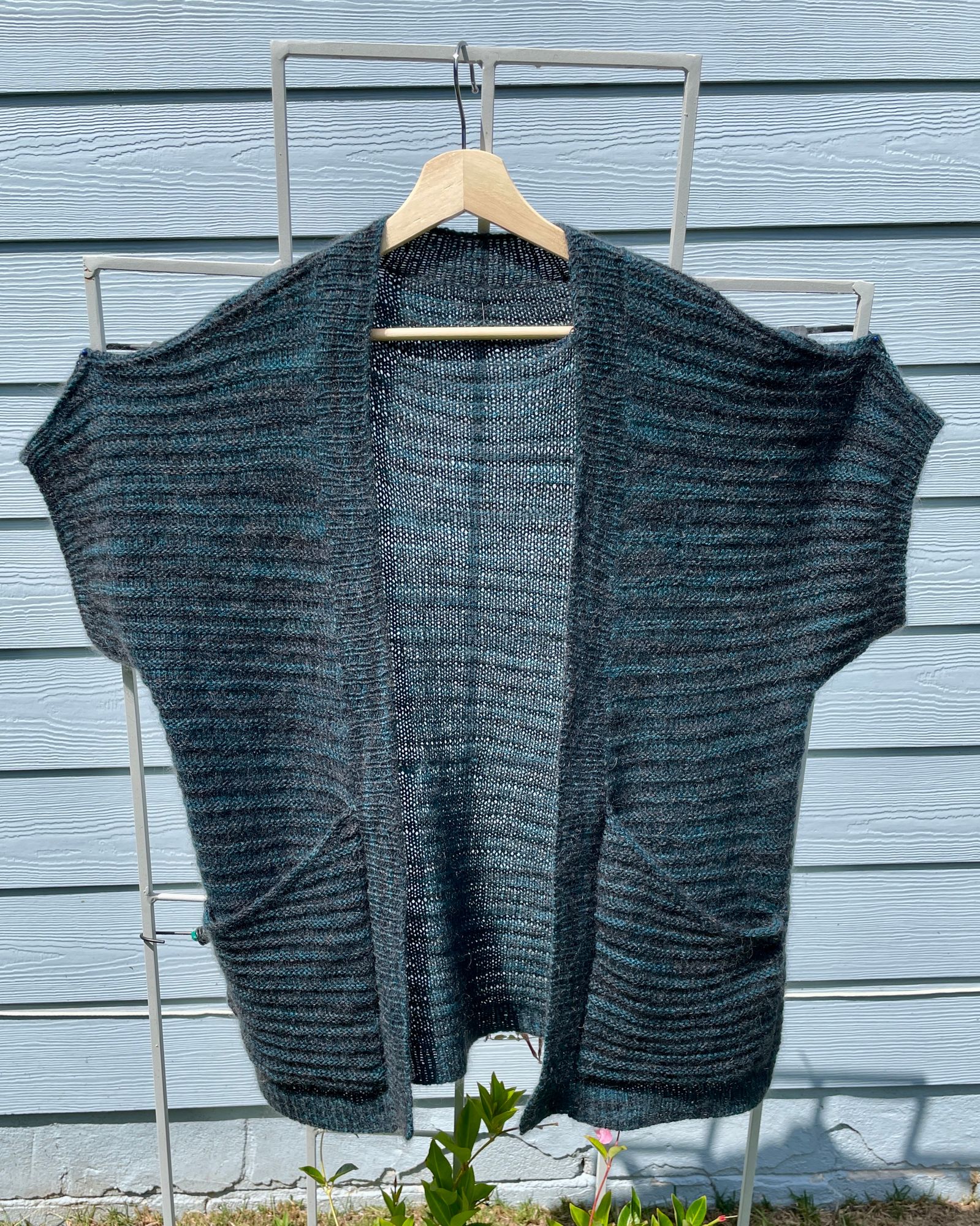 A sort of short-sleeved handknitted cardigan. The "sleeves" are formed like a drop-shoulder sweater, with ribbed hems. The yarn is a variegated blue/green/dark blue laceweight yarn combined with a very dark grey mohair yarn. The fabric is a little sheer. There are deep pockets at the bottom.