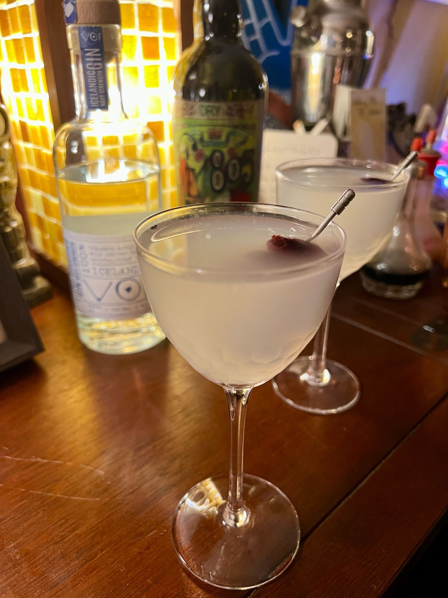 Two cloudy Icelandic Martinis atop our bar, with the gin and vermouth bottles behind them, the olive pick garnish submerged in the drinks. Backlit by our mahogany-framed glass tile bar lamp.