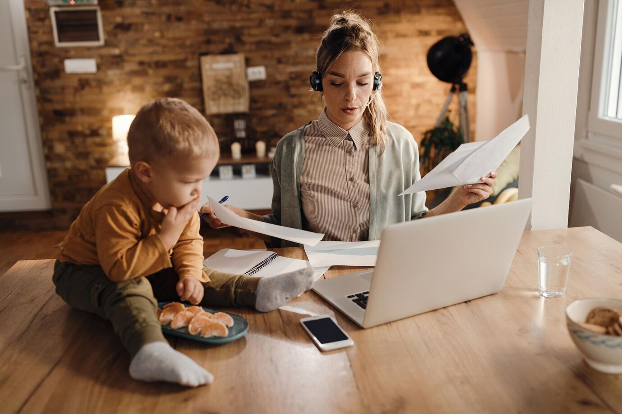 Imagem de uma mãe trabalhando no computador, com um headset e segurando papéis, enquanto o filho está sentado em cima da mesa, ao lado do notebook e celular.