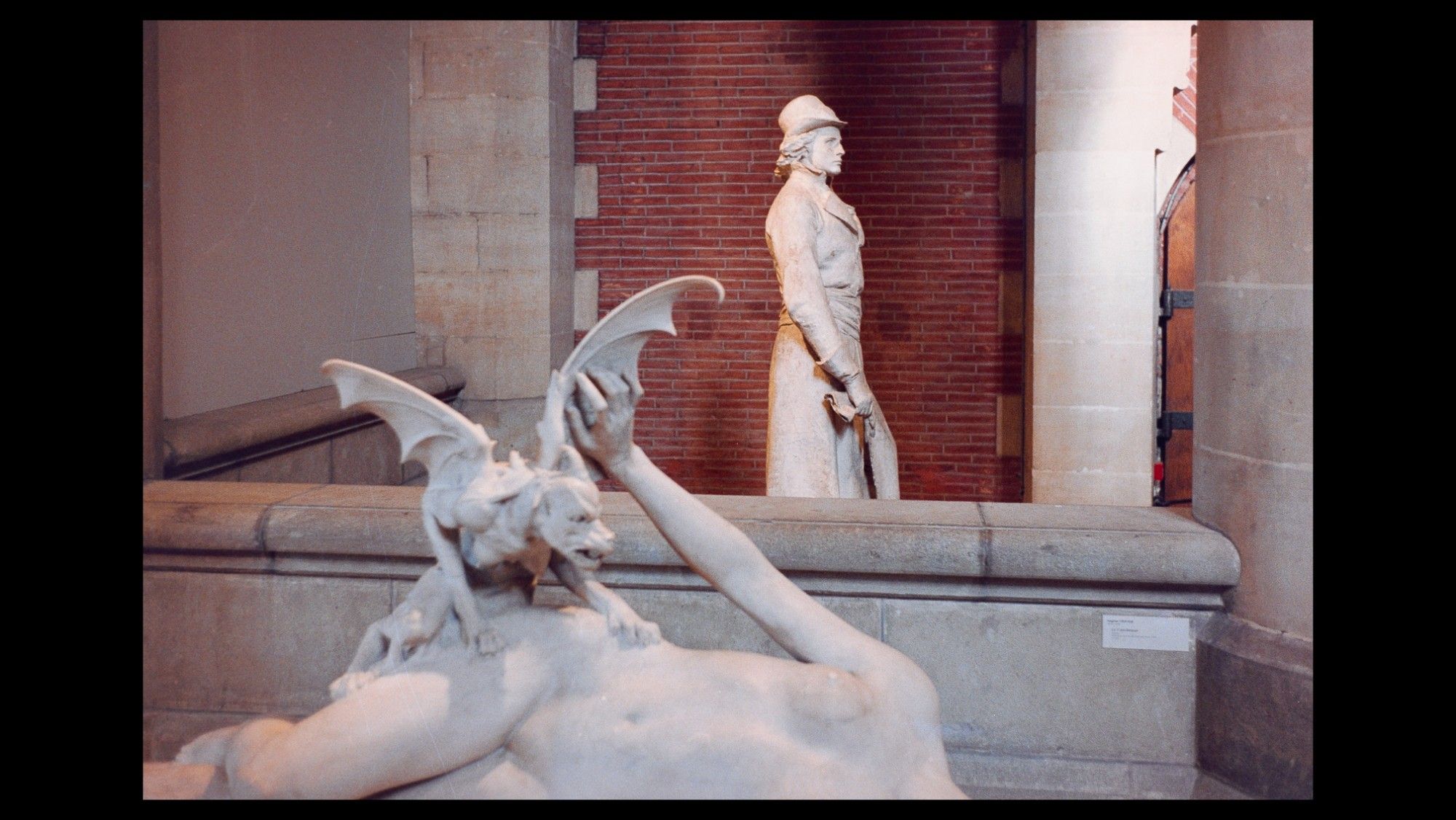 Museum interior of stone- and brick-work with marble statuary. In the foreground, a dog-headed, bat-winged imp torments a nude maiden. Behind a low wall, a slender elegantly dressed young man resolutely ignores them.