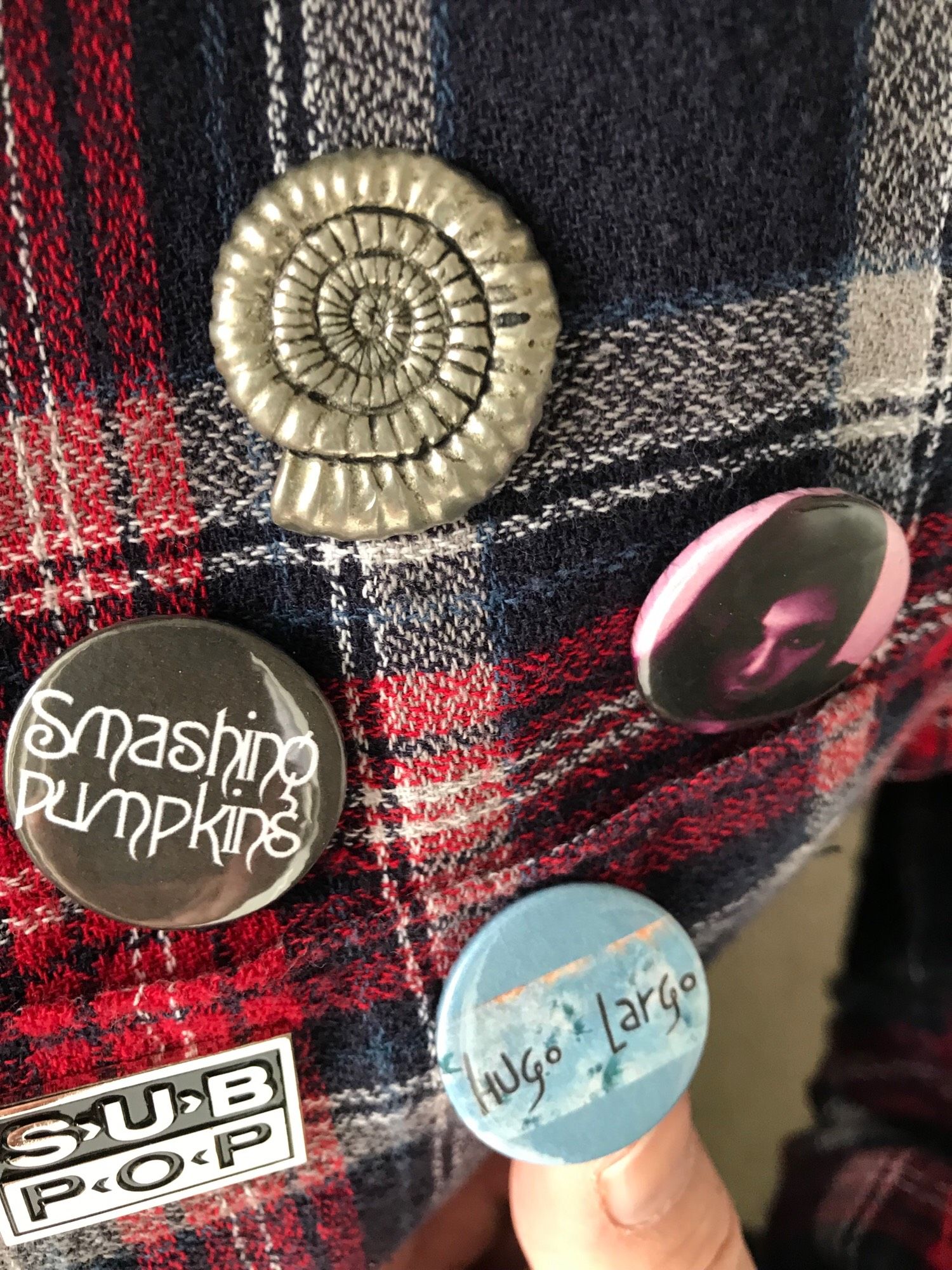 Badges for Nicola Griffith’s Ammonite, Izumi Suzuki, Smashing Pumpkins, Hugo Largo and Sub Pop records on a plaid shirt