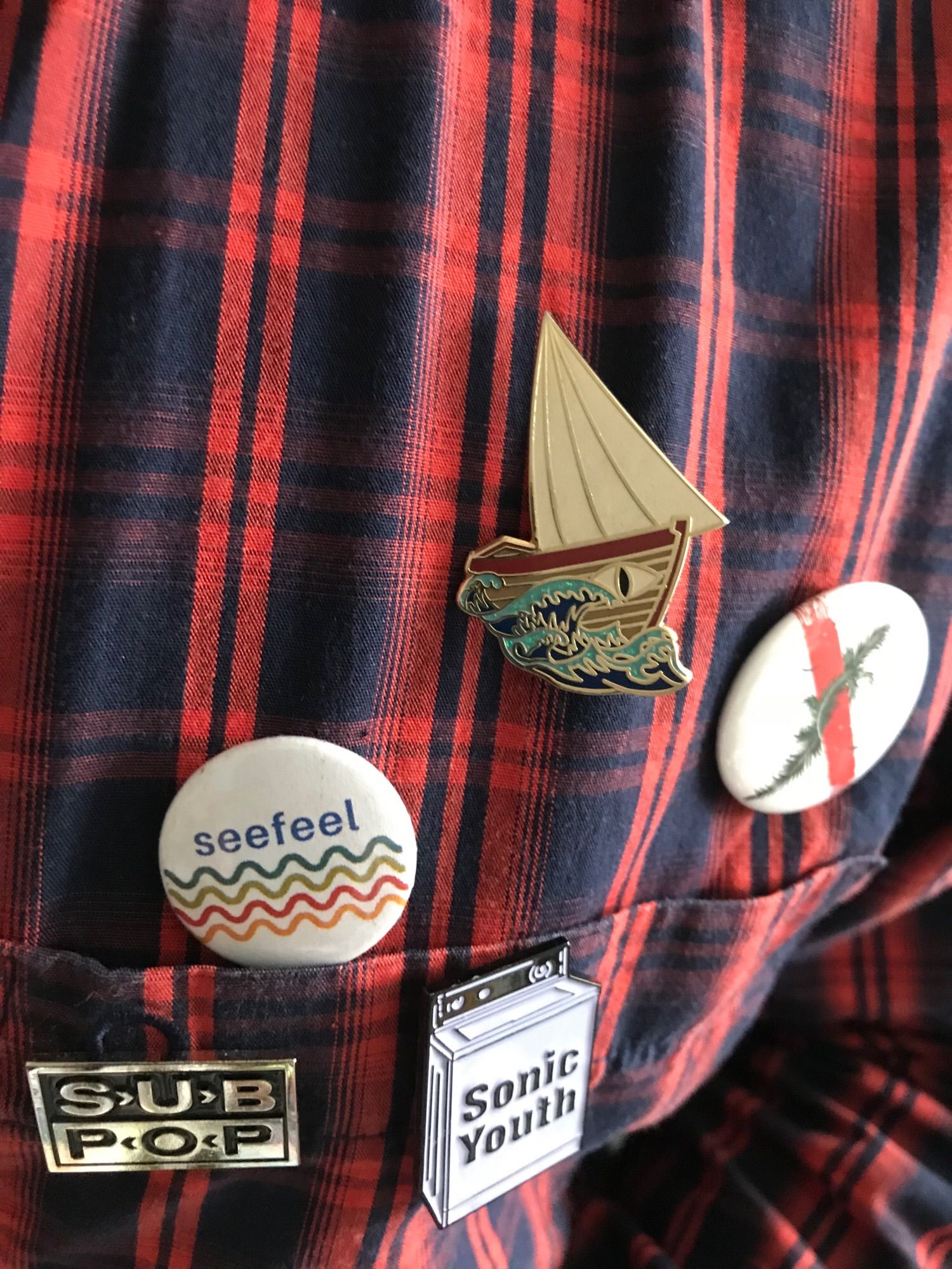 Badges for Ursula Le Guin’s Lookfar, Jeff VanderMeer’s Area X, Seefeel, Sonic Youth and Sub Pop on a plaid shirt