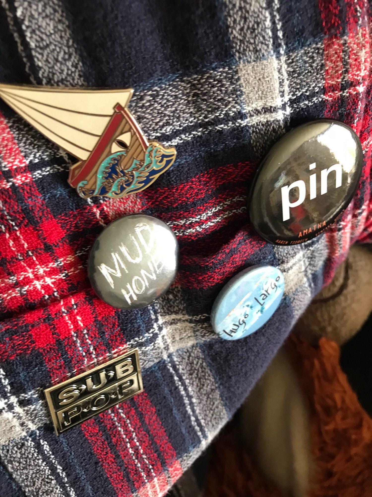 Badges for Ursula Le Guin’s Lookfar from Earthsea, Karin Tidbeck’s Amatka, Mudhoney, Hugo Largo and Sub Pop records on a plaid shirt