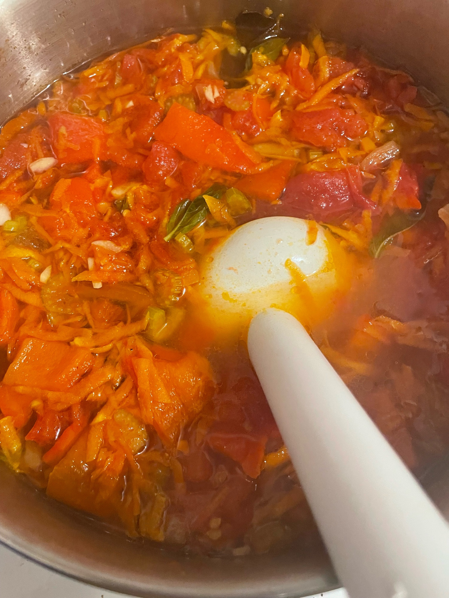 A mix of grated carrots, chunks of celery, red pepper, tomatoes, garlic and vegetable broth, about to be blended with an immersion blender. 