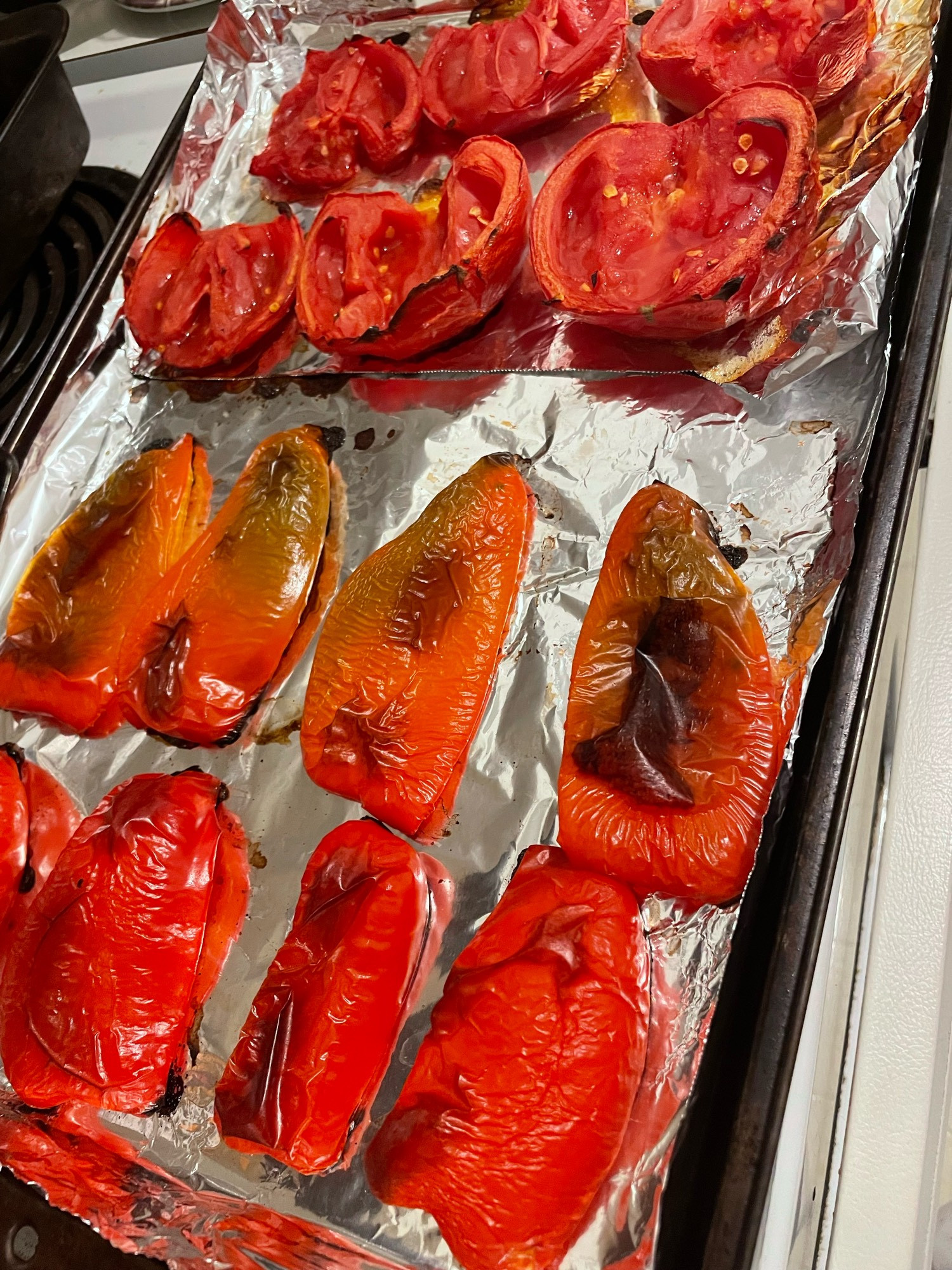 A foil-lined baking sheet with rows of roasted red peppers and roasted tomatoes cooling down so they can be peeled and put in the pot. 