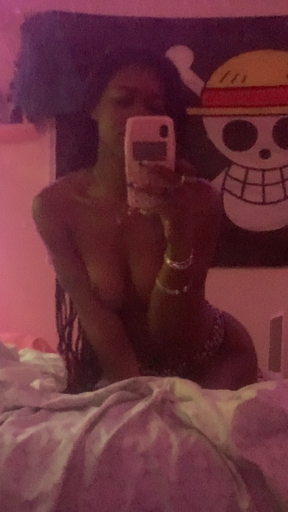 Nude red tinted mirror selfie kneeling on grey sheets leaning forward so my silhouette is outlined bt you can barely see my body. Long braids hang to my side. Cleavage visible as my small tits hang jst a bit. One piece jolly Roger skull is in view over my right shoulder my phone case is a pink Nokia 